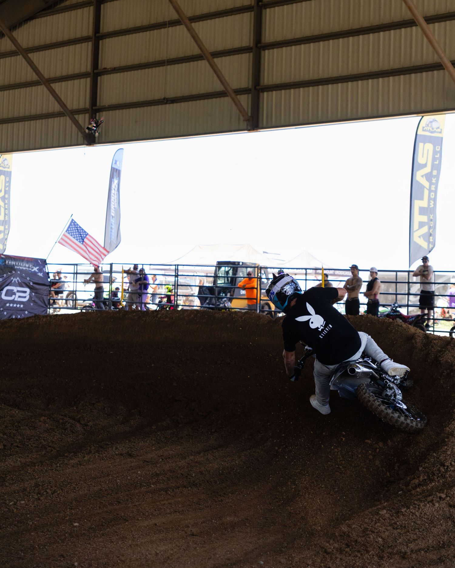 Motocross rider leans into a dirt berm corner inside an indoor supercross track with spectators.