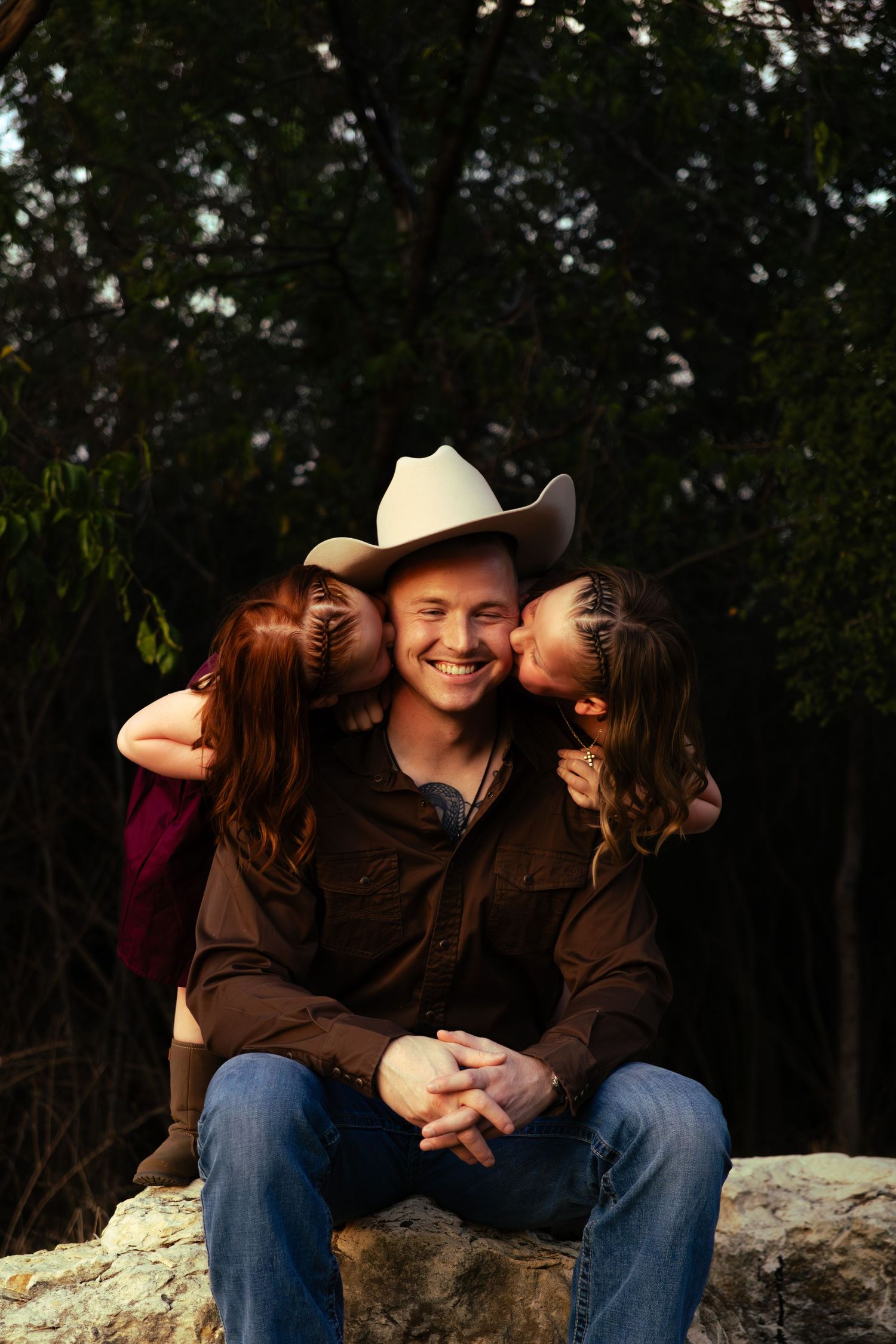 Smiling man in cowboy hat sitting on rocks while two girls kiss his cheeks outdoors.