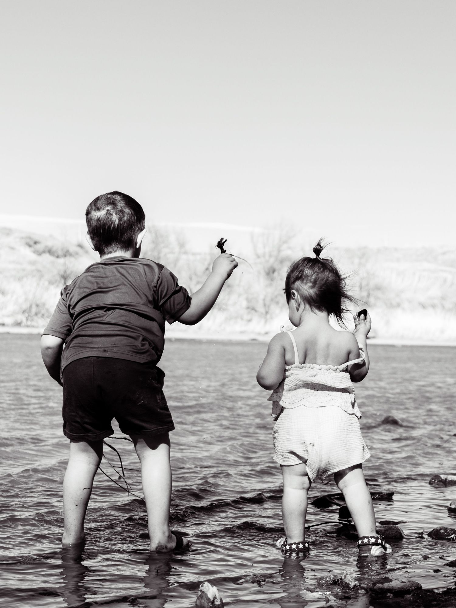 Two young children wading in rocky shallow water near a lake shore in black and white photo.