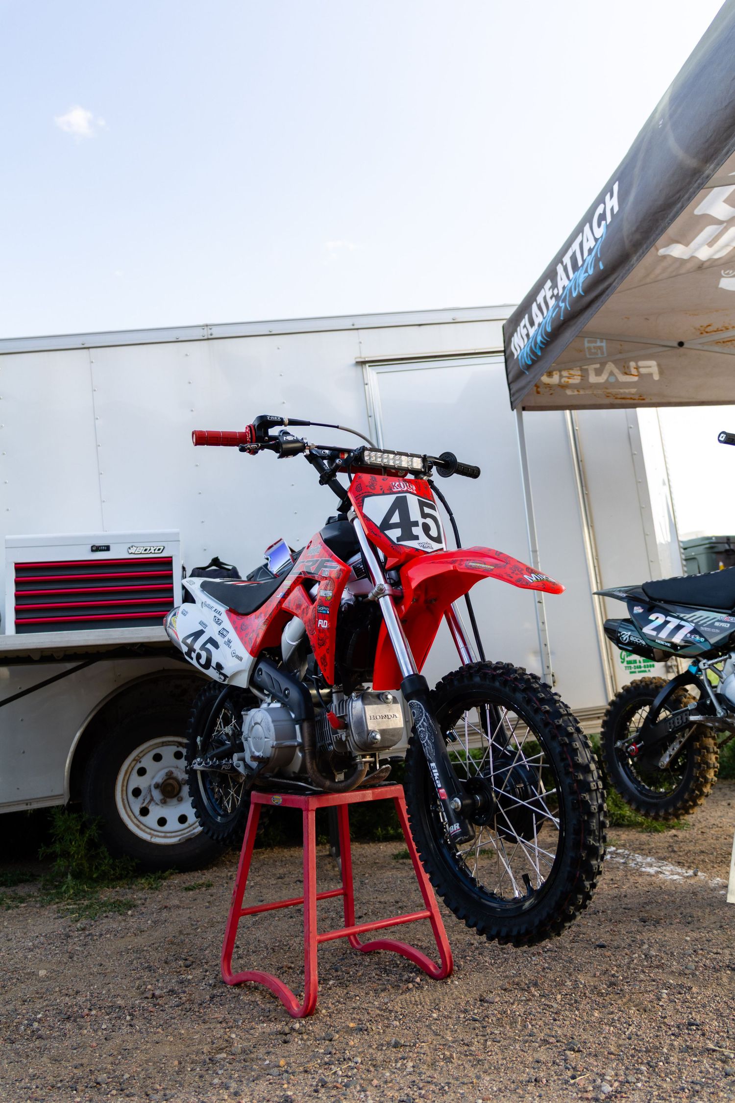 Red dirt bike number 45 on a stand outside a trailer at an outdoor motocross racing event.