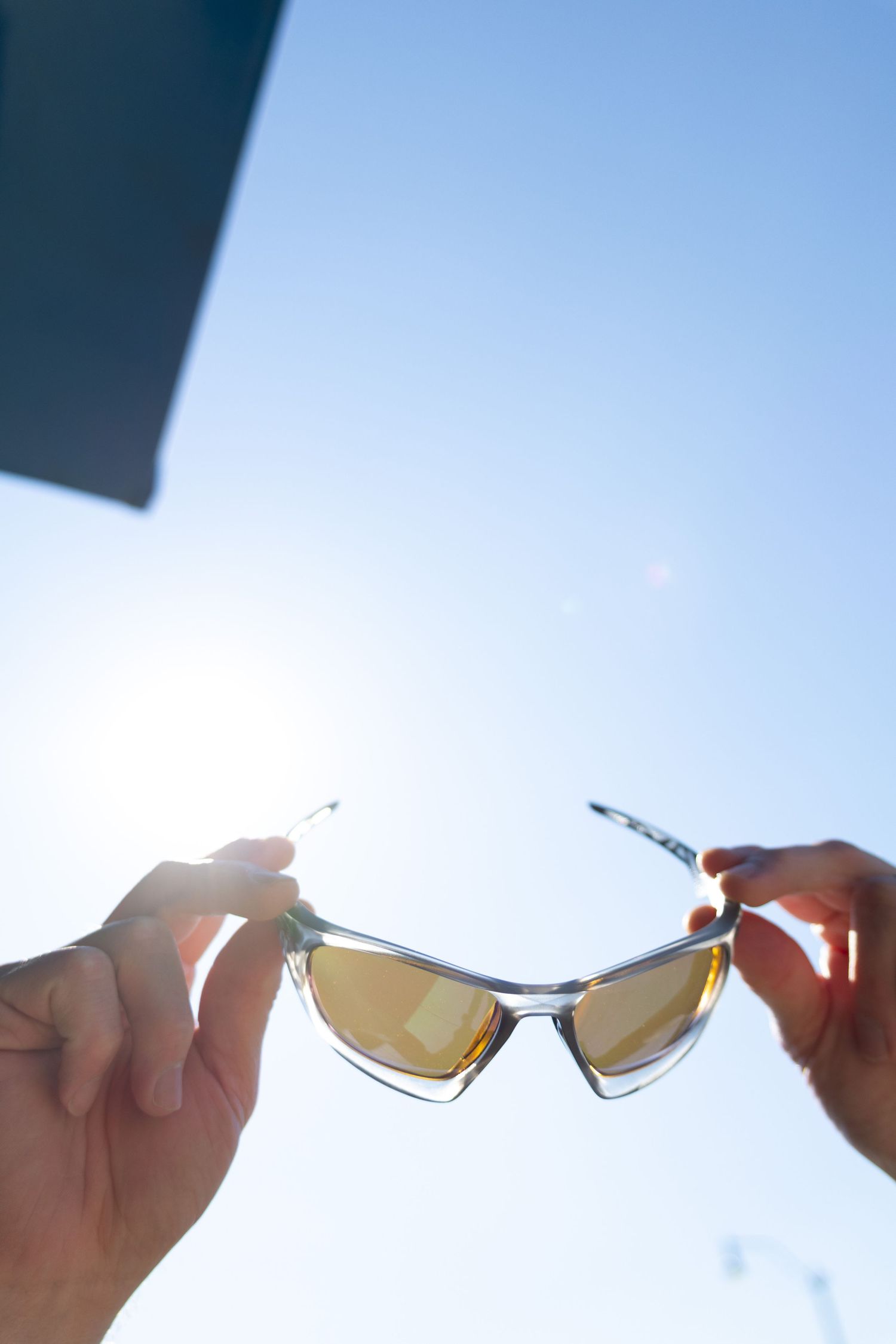 Hands holding sport sunglasses up toward a bright blue sunny sky with lens flare in color photo.