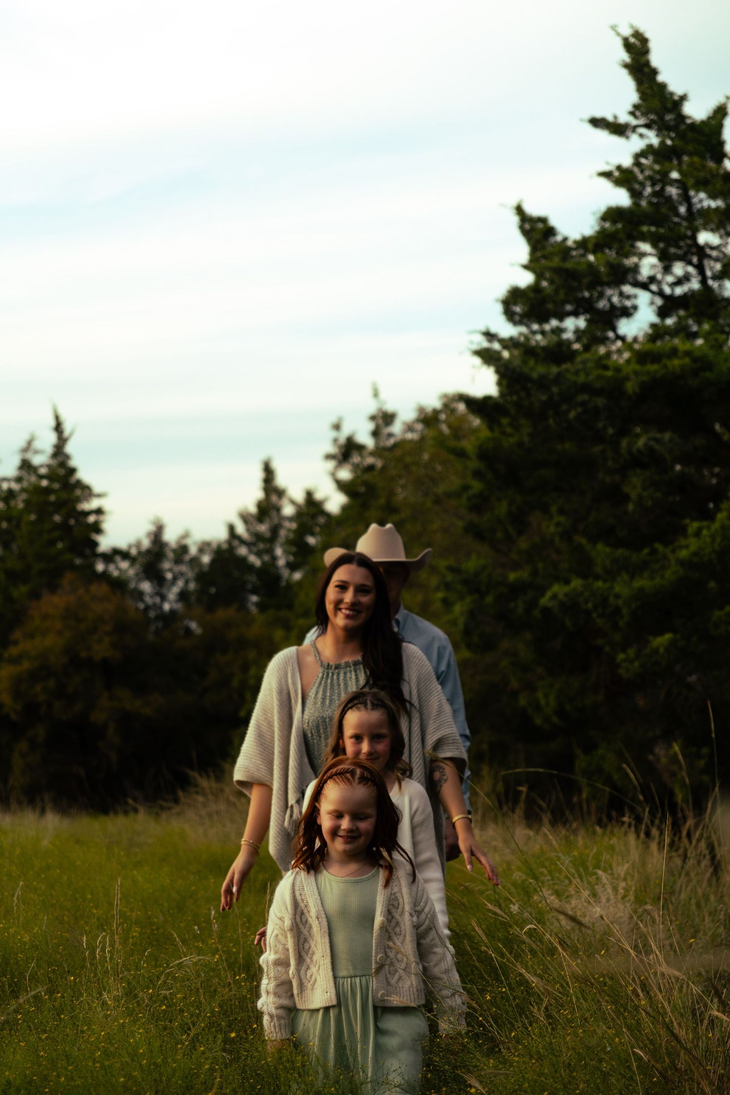 Mother and two daughters walking through a grassy field with tall evergreen trees in the background.