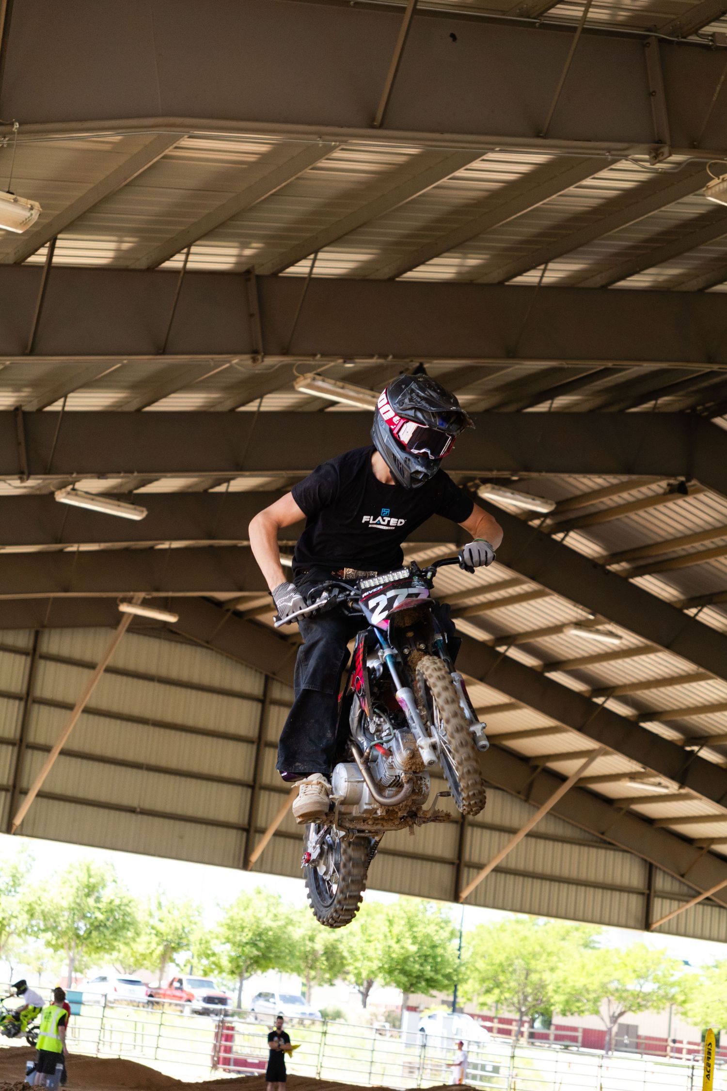 Motocross rider in black gear launching off a dirt jump inside a covered arena during a supercross event.