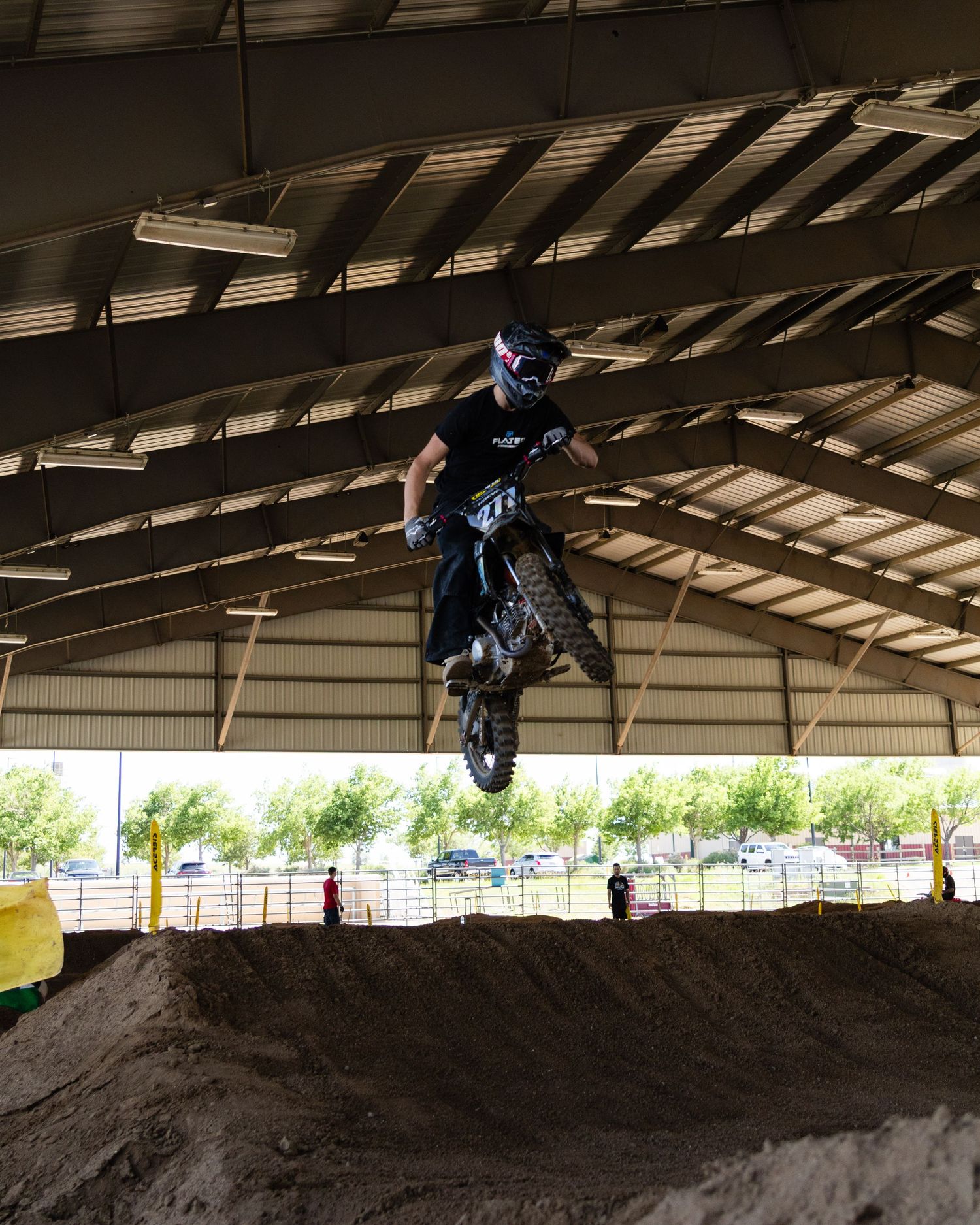 Dirt bike rider in all black gear soars over large dirt mound at indoor motocross event.