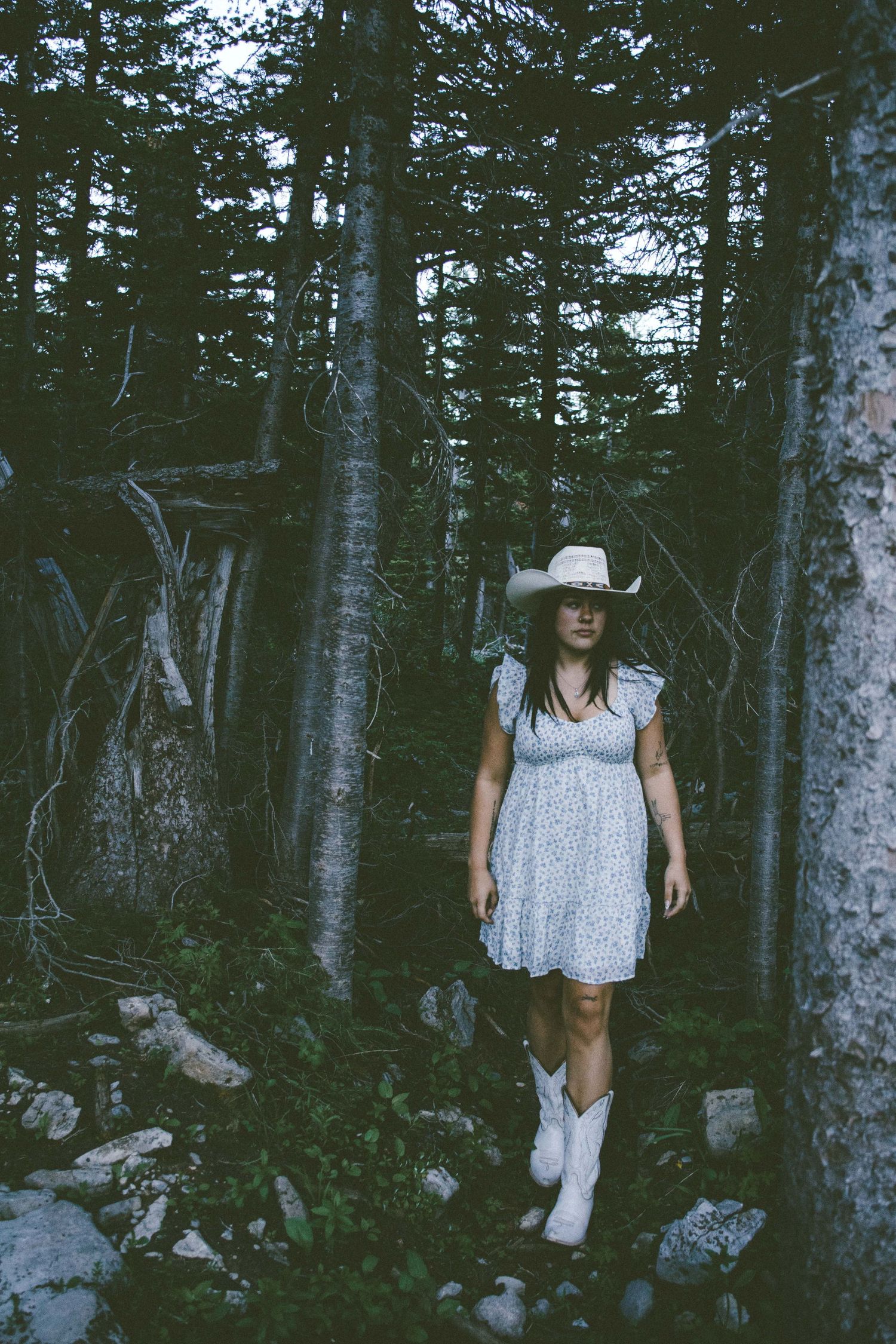 Woman in white cowboy hat, floral dress, and white boots walking through a dark forest trail.