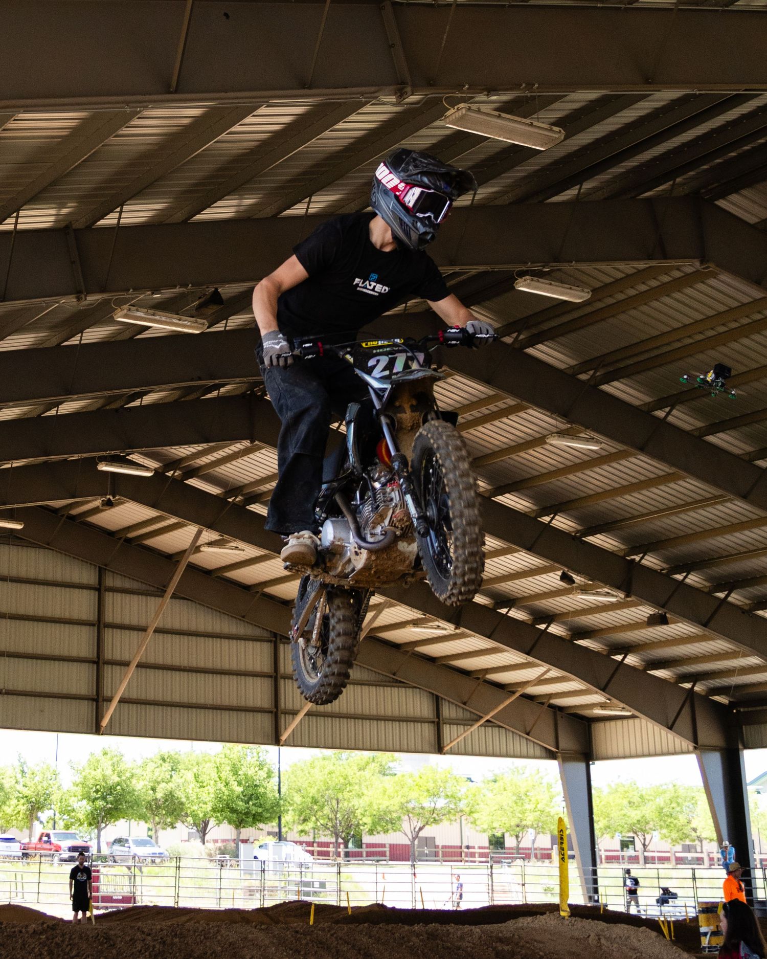 Motocross rider performing big jump on dark dirt bike inside covered arena with spectators.