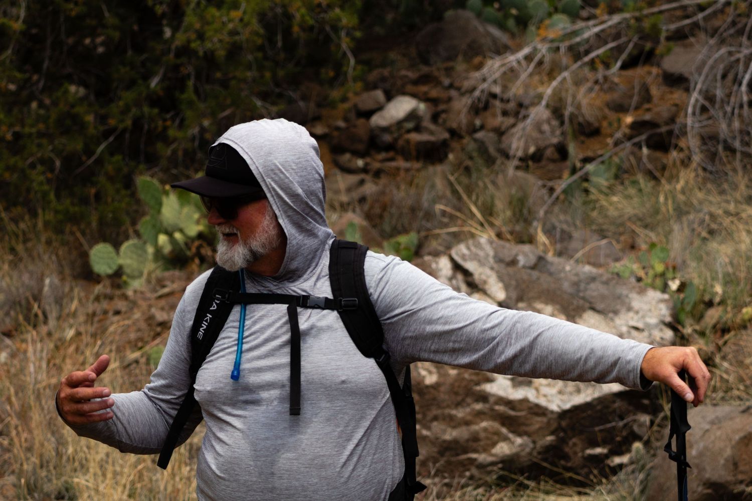 Man in gray hoodie with backpack and hiking pole navigating rocky terrain on a trail.