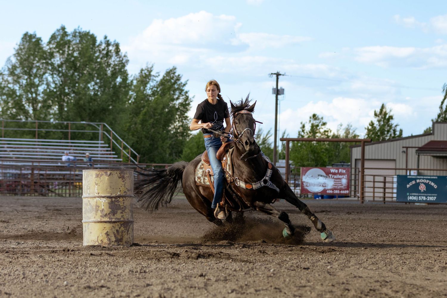 Barrel racer on dark horse making sharp turn around barrel at outdoor rodeo arena during evening competition.