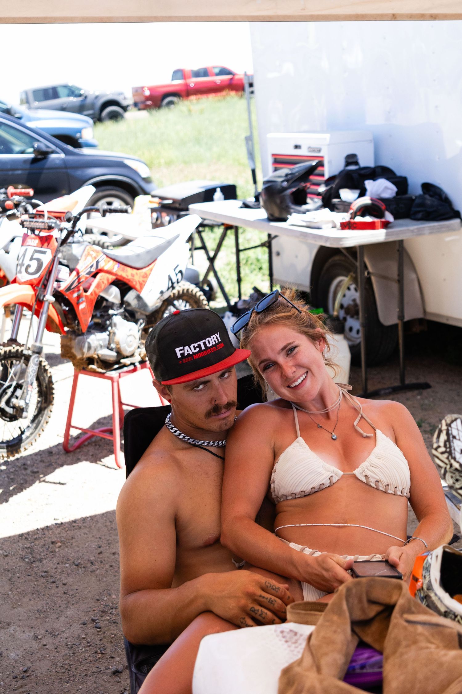 Young couple relaxing together at a motocross racing event with dirt bikes and trailer in background.