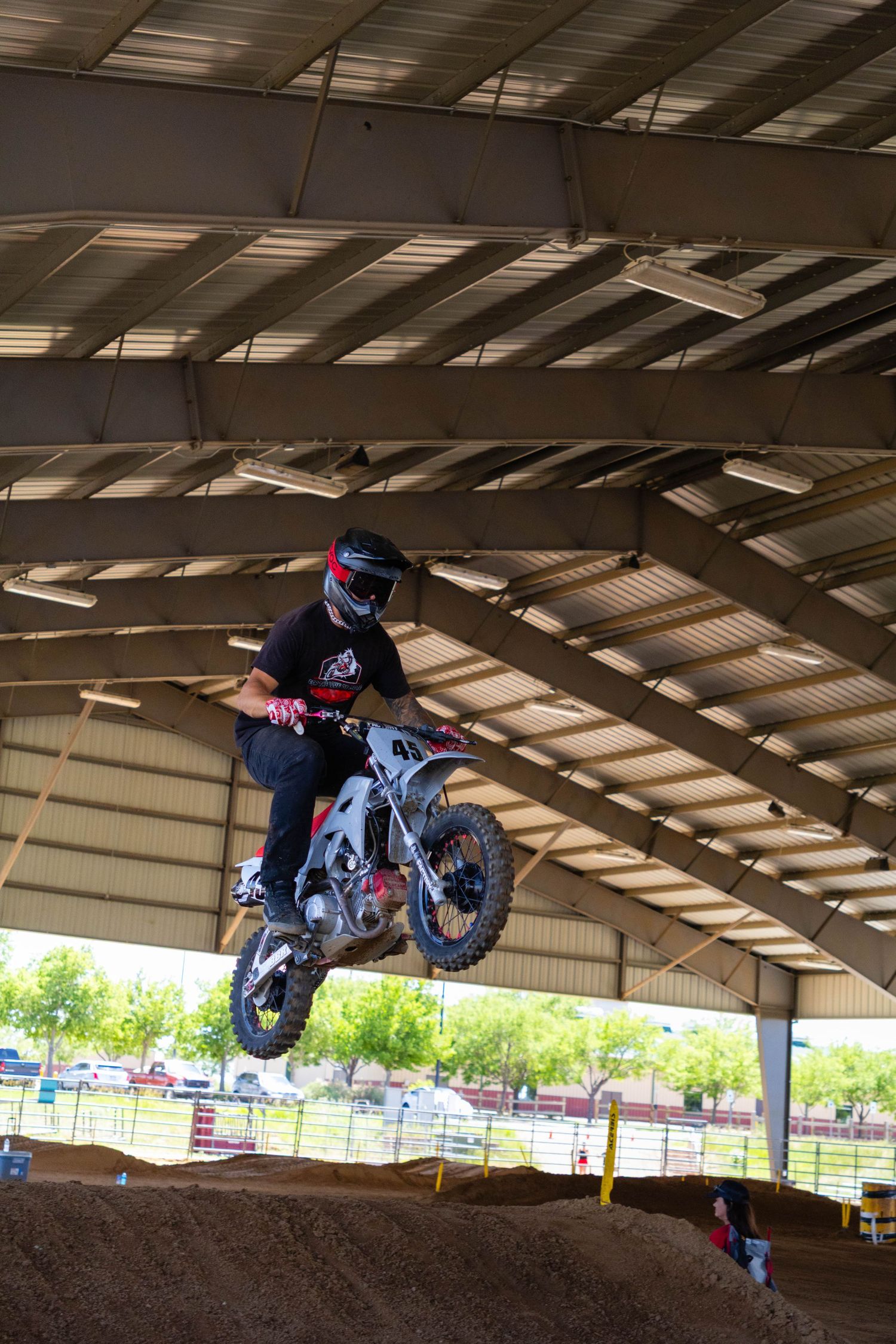 Motocross rider launching off a dirt jump inside a large covered indoor arena during a race.