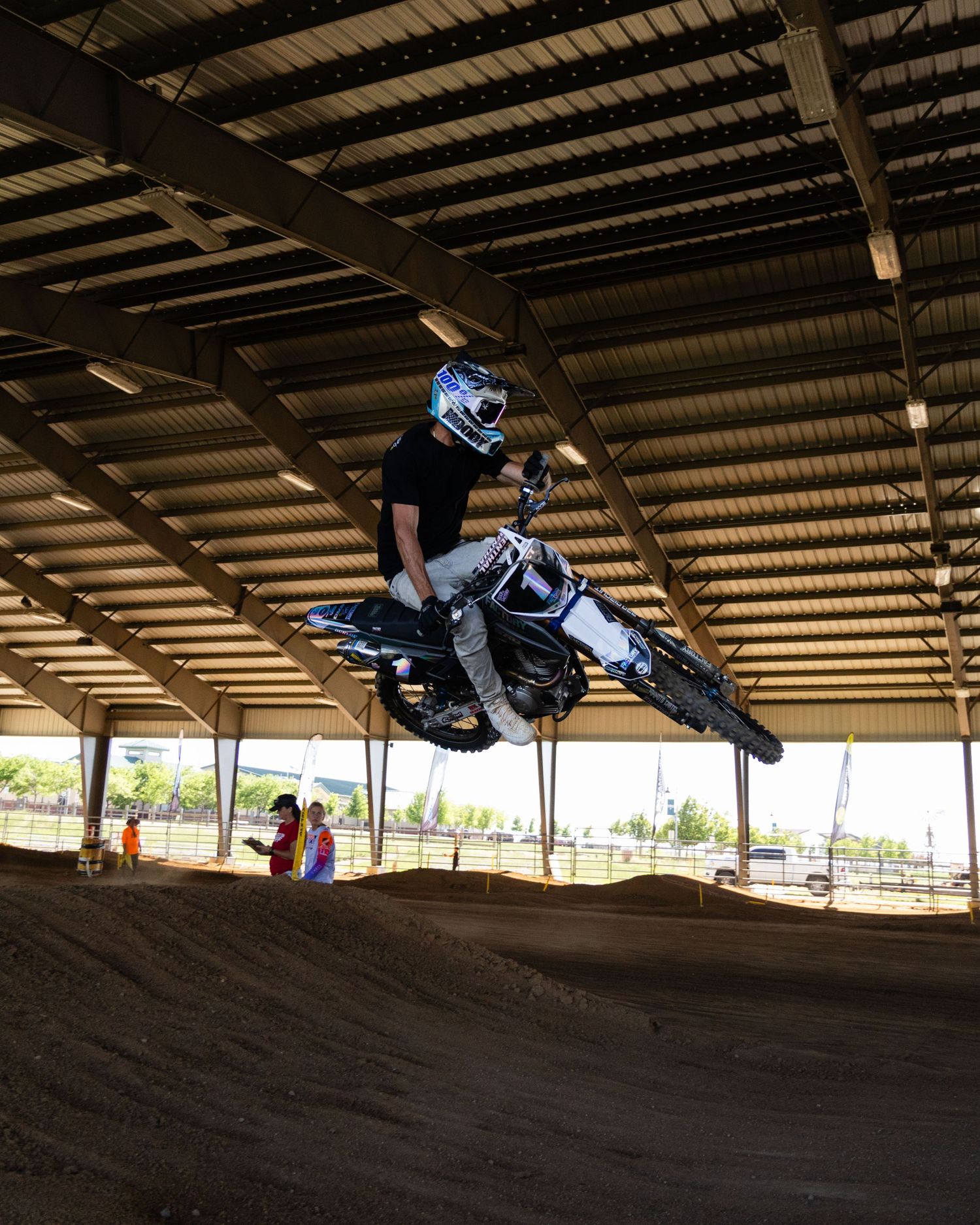 Motocross athlete soaring over a dirt jump inside a covered arena during practice.
