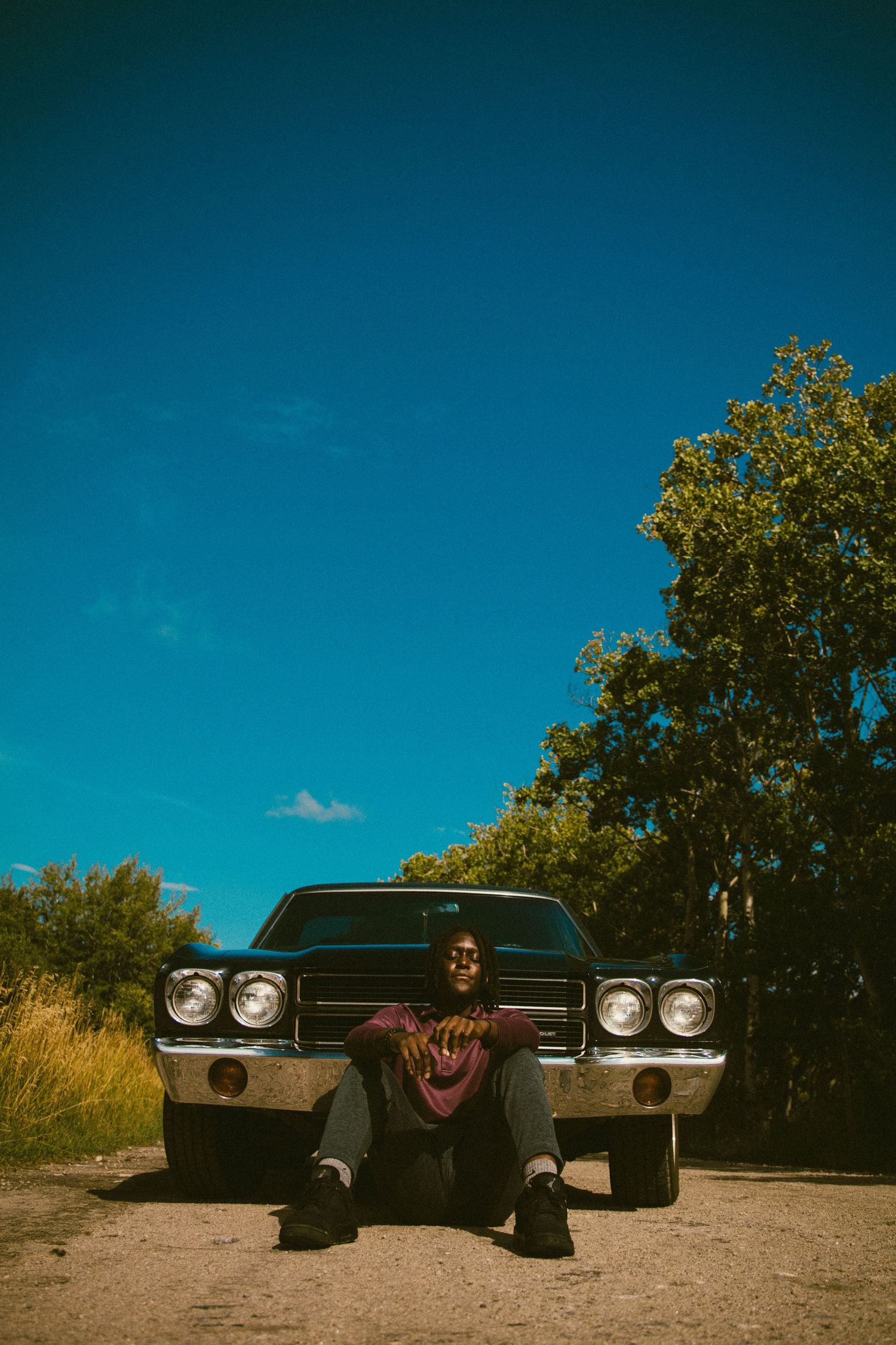 Young man sitting in front of a classic black muscle car on a dirt road under a vivid blue sky.
