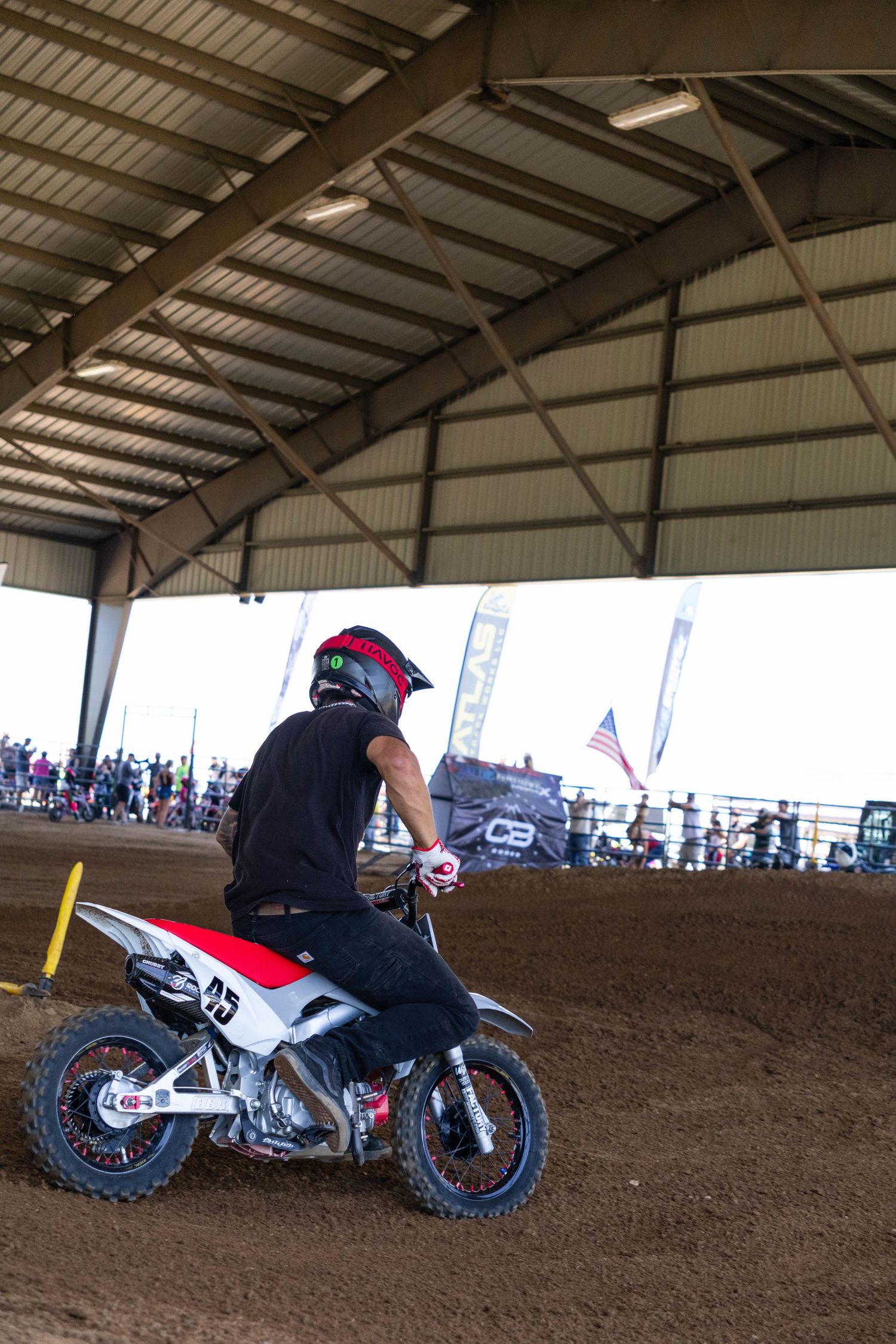 Motocross rider on red and white dirt bike navigating indoor dirt track with crowd watching.