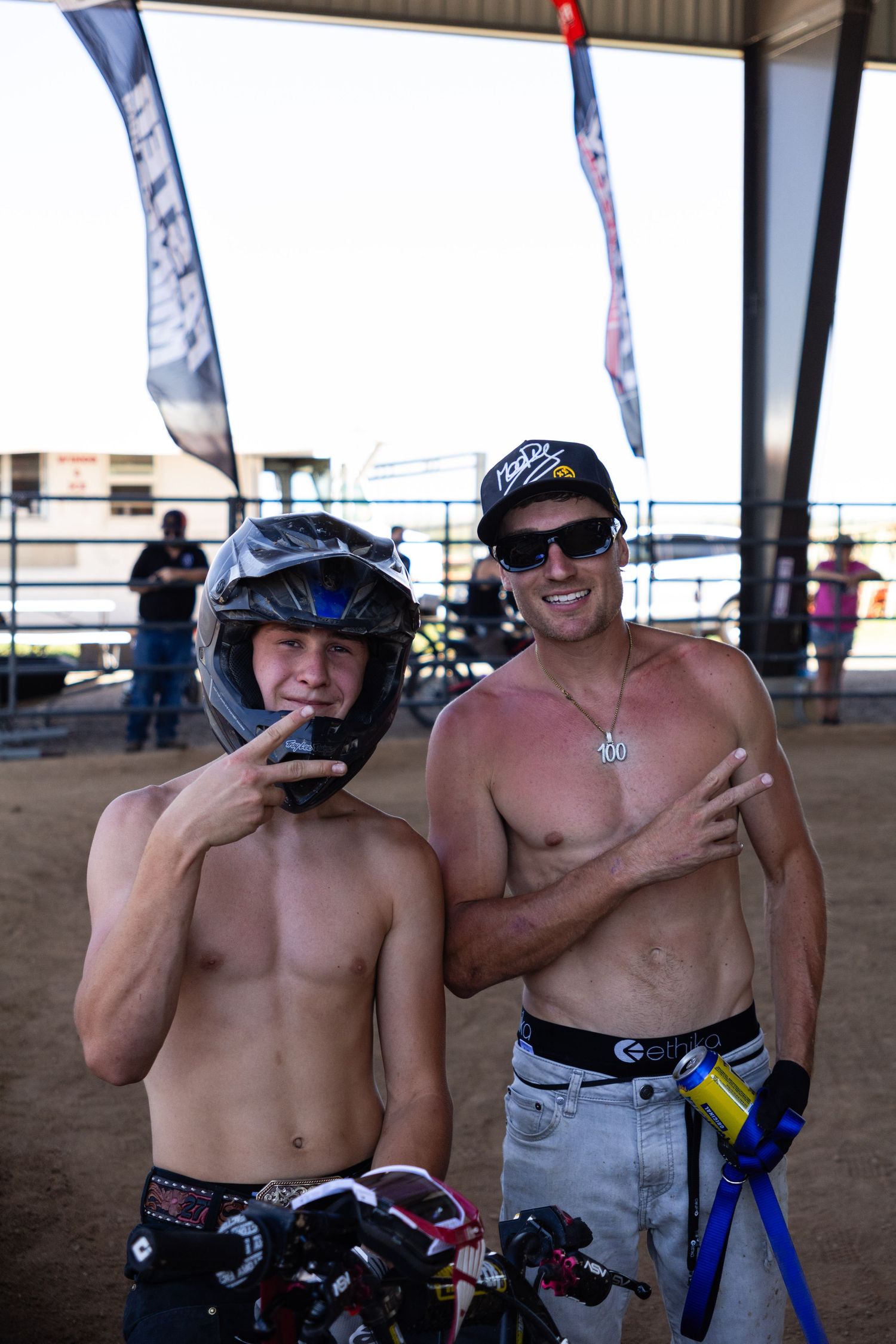 Two shirtless motocross riders pose together at an outdoor dirt arena, one wearing a helmet.