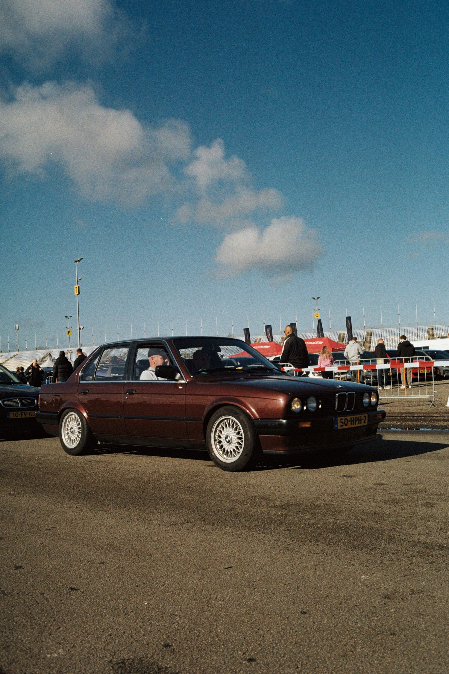 Classic burgundy BMW E30 sedan parked at an outdoor car meet under blue sky.