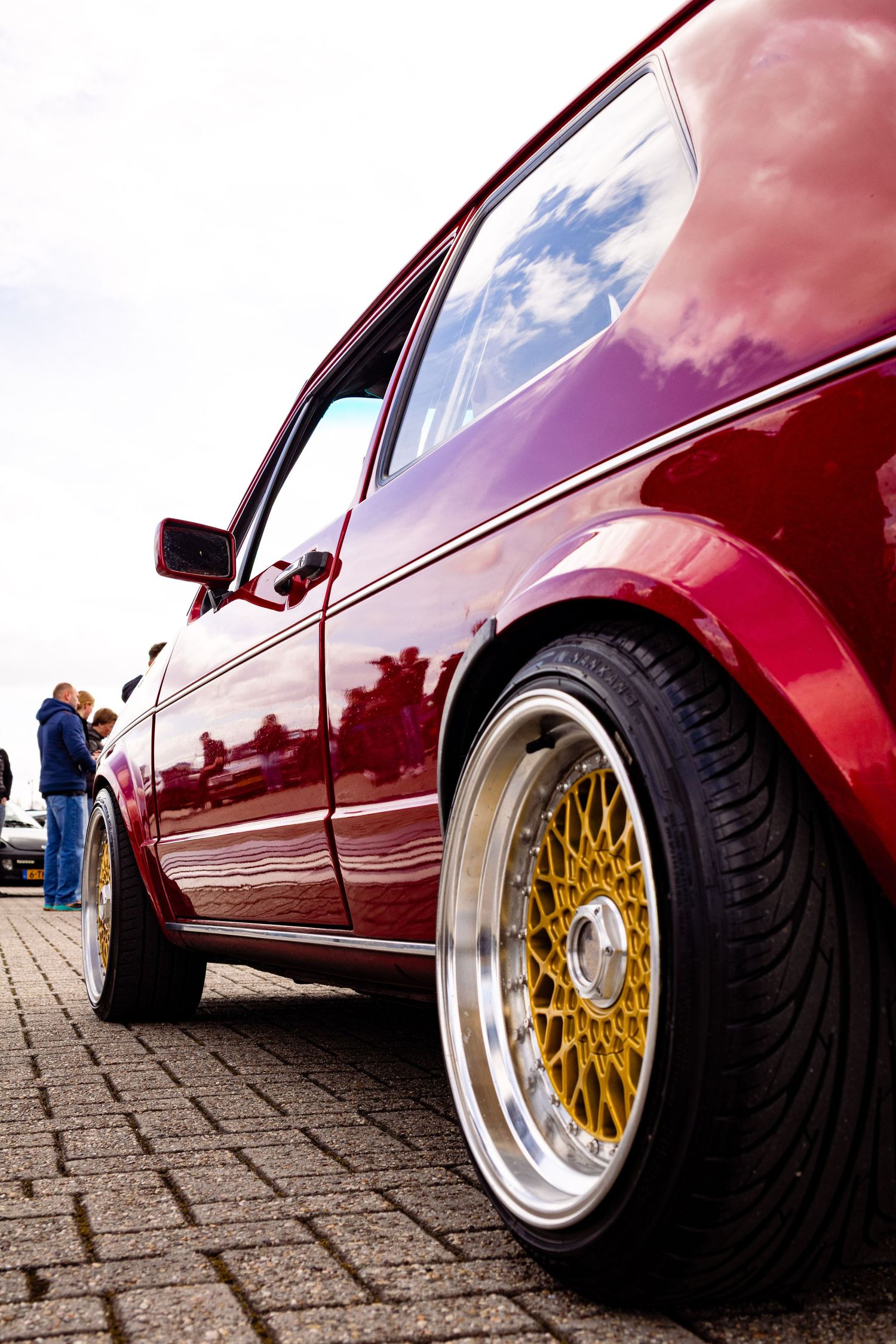 Low angle view of a shiny red car with gold BBS wheels on cobblestone pavement.