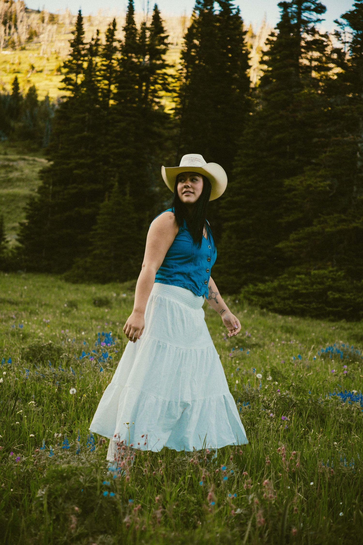 Woman in blue top, white skirt, and cowboy hat walking through a wildflower meadow with pine trees.