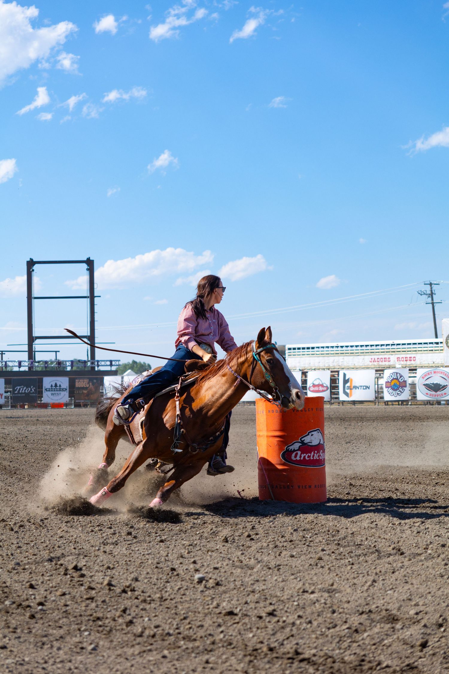 Female barrel racer on a chestnut horse rounding an orange barrel at a sunny rodeo event.