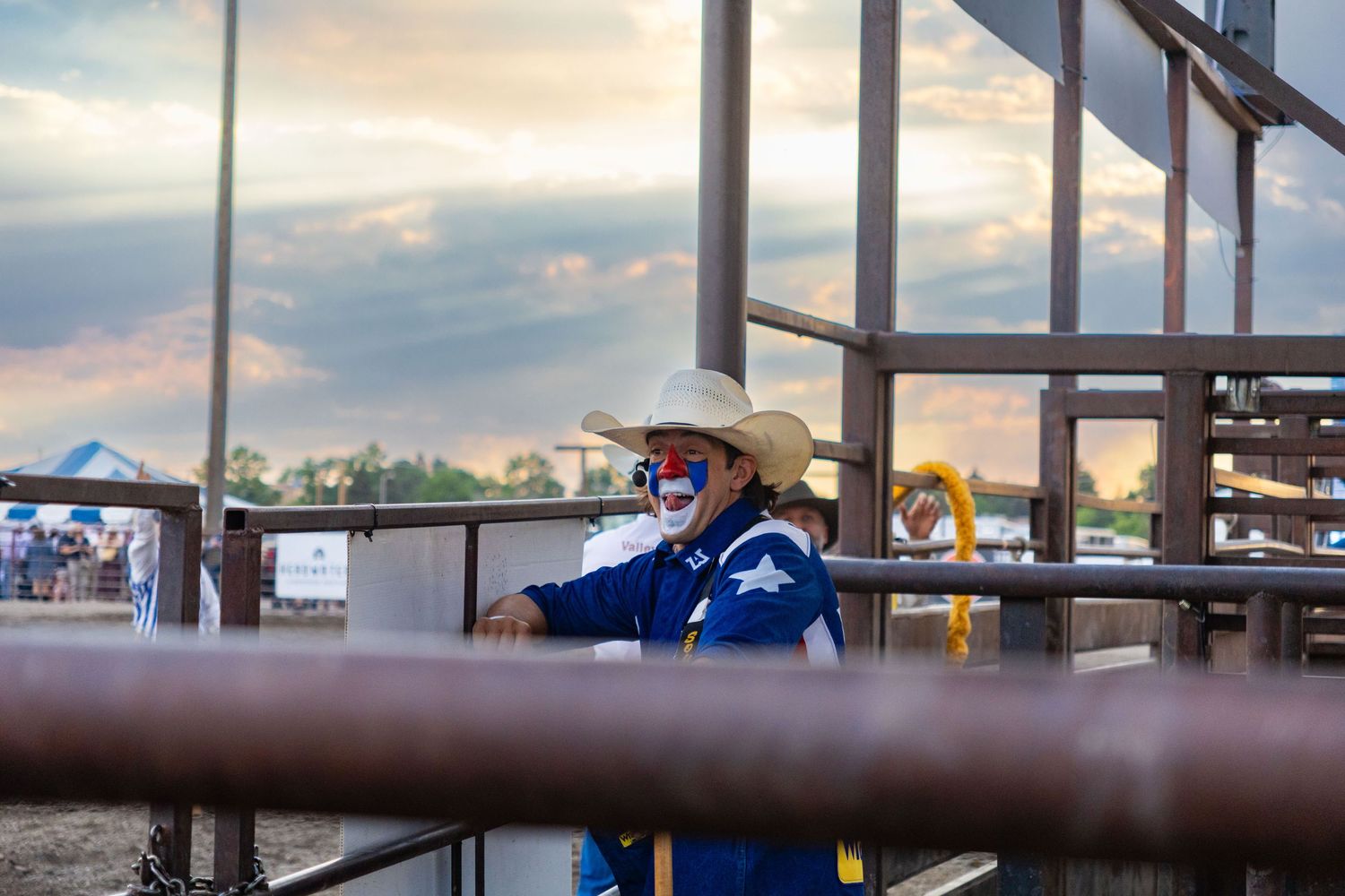 A rodeo clown in a blue costume and face paint stands at the arena fence at sunset.