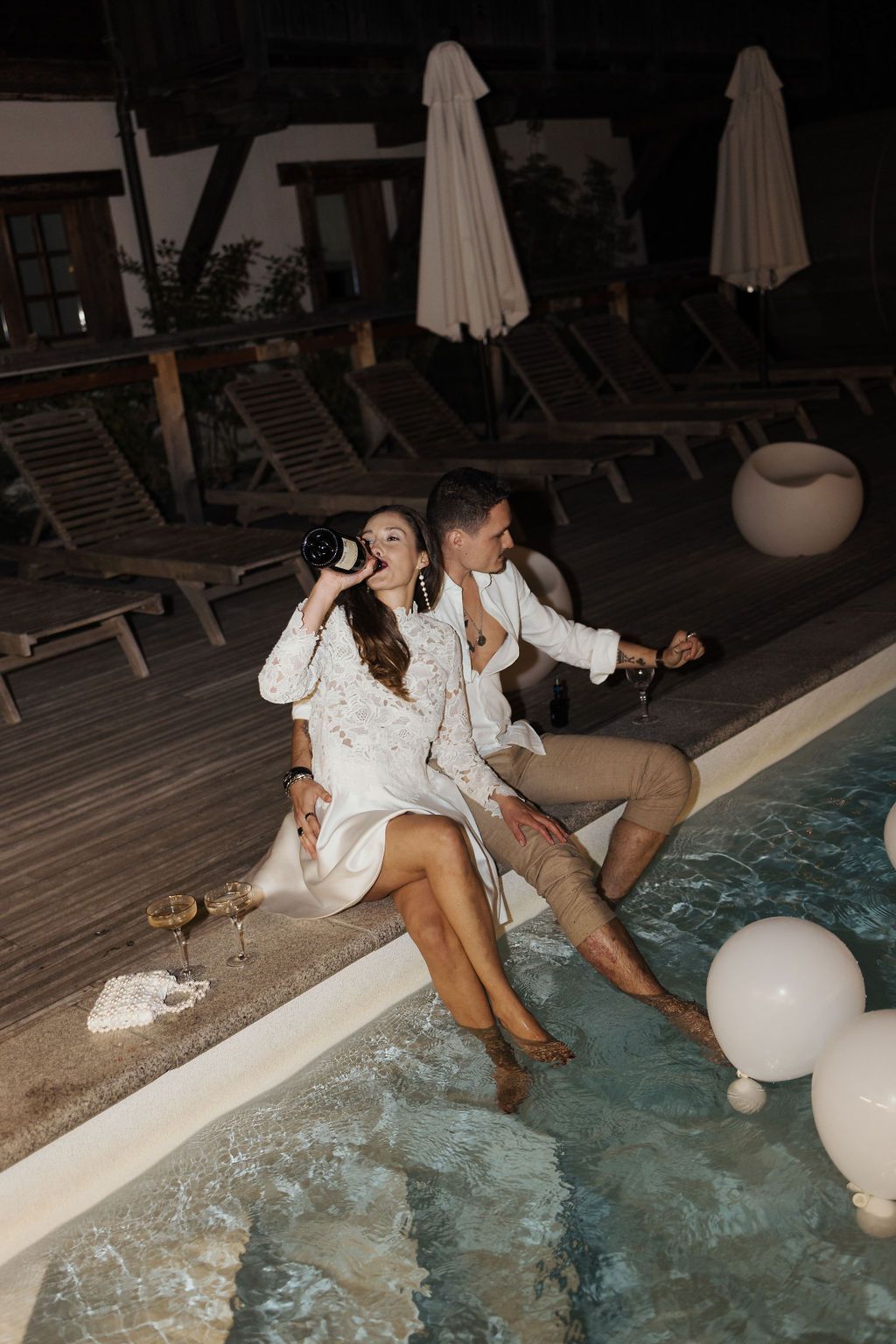 Newlywed couple laughing and drinking champagne by illuminated pool at night.