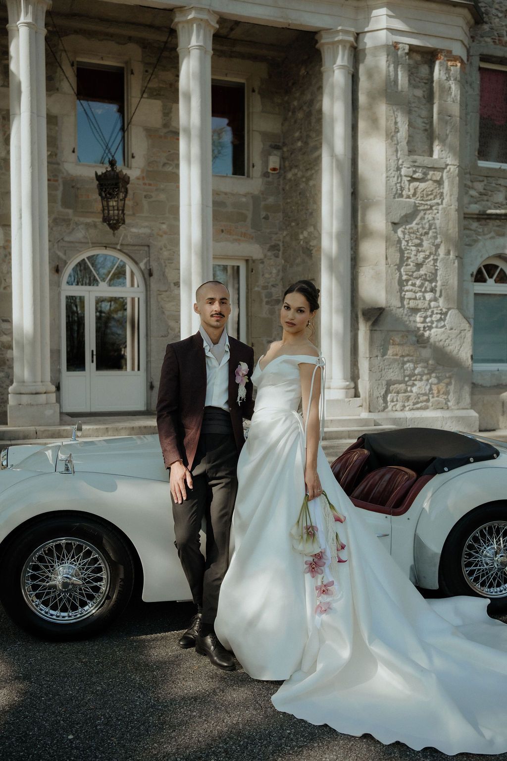 Bride and groom posing beside vintage white convertible car in front of stone manor house.