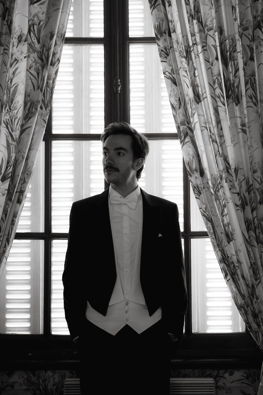 Dapper groom in black tuxedo and white waistcoat stands thoughtfully before large window with floral curtains.