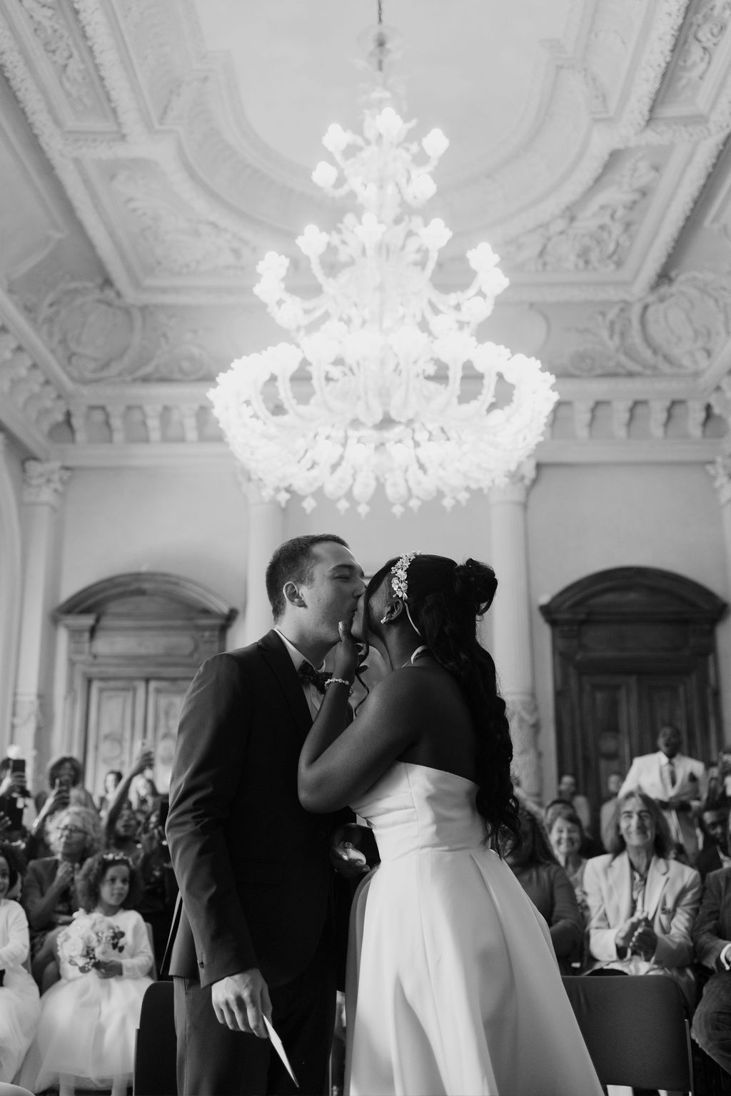 Black and white photo of interracial couple sharing first kiss at wedding ceremony under grand chandelier.
