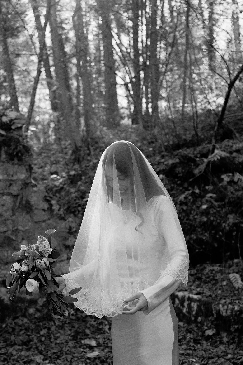 Black and white portrait of bride in satin gown holding bouquet with lace veil draped over head in forest setting.