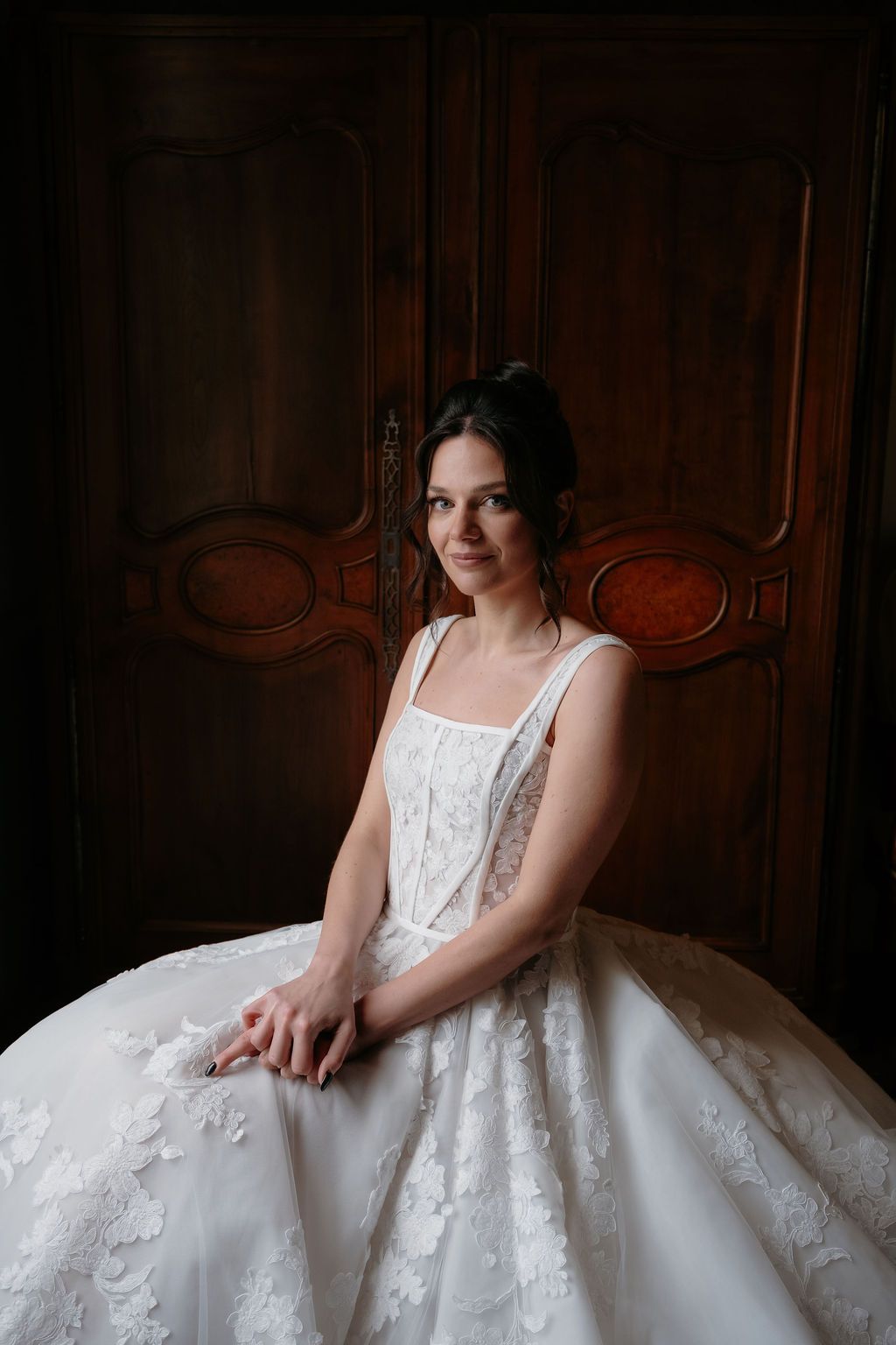 Bride in lace floral ballgown sitting serenely before antique wooden armoire in dim lighting.