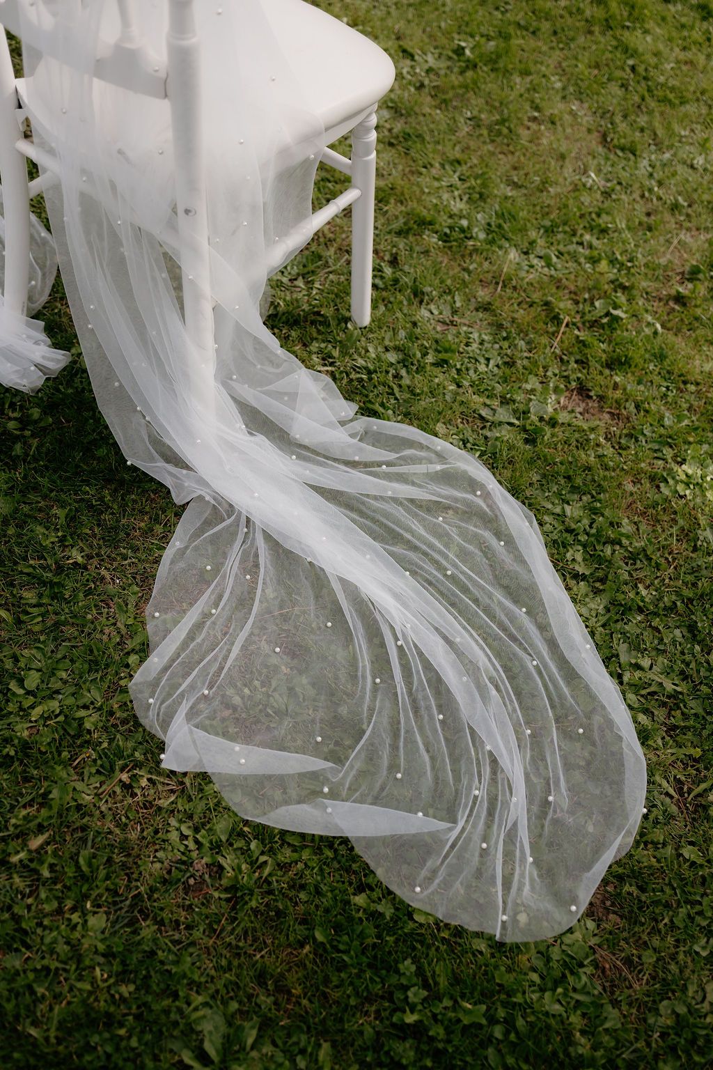 White pearl-studded wedding veil draped elegantly over a chiavari chair on green grass.