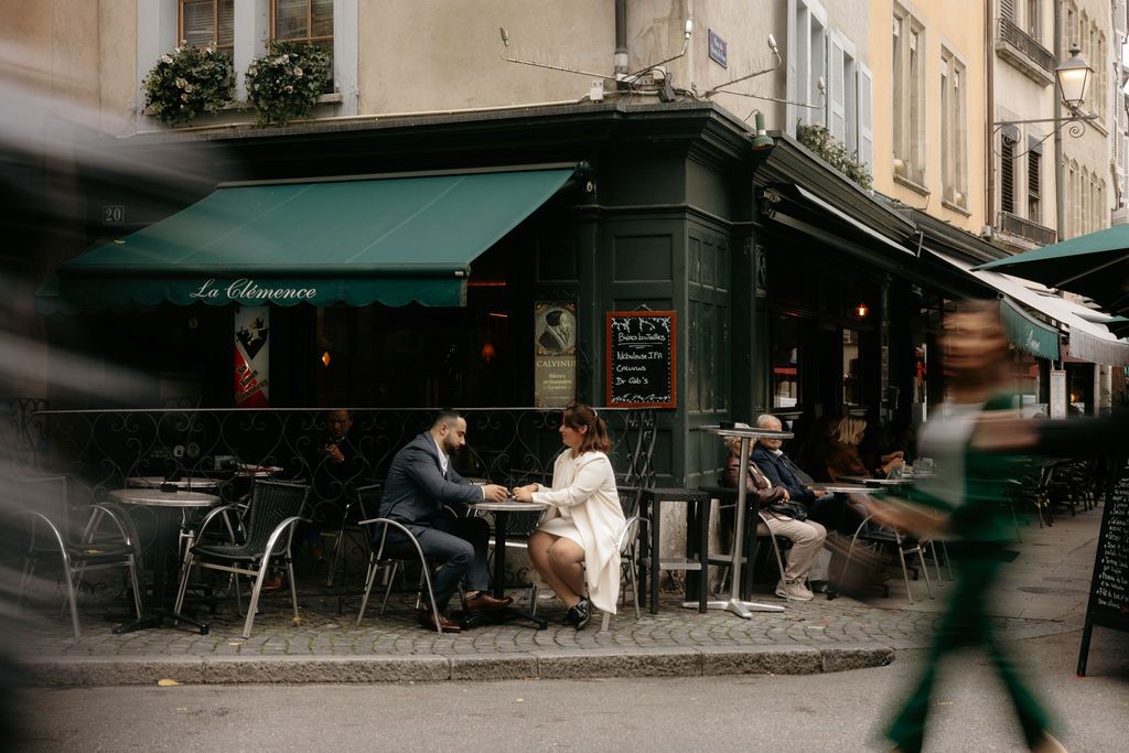 Couple seated at an outdoor Parisian café terrace with green awning, surrounded by passing pedestrians.