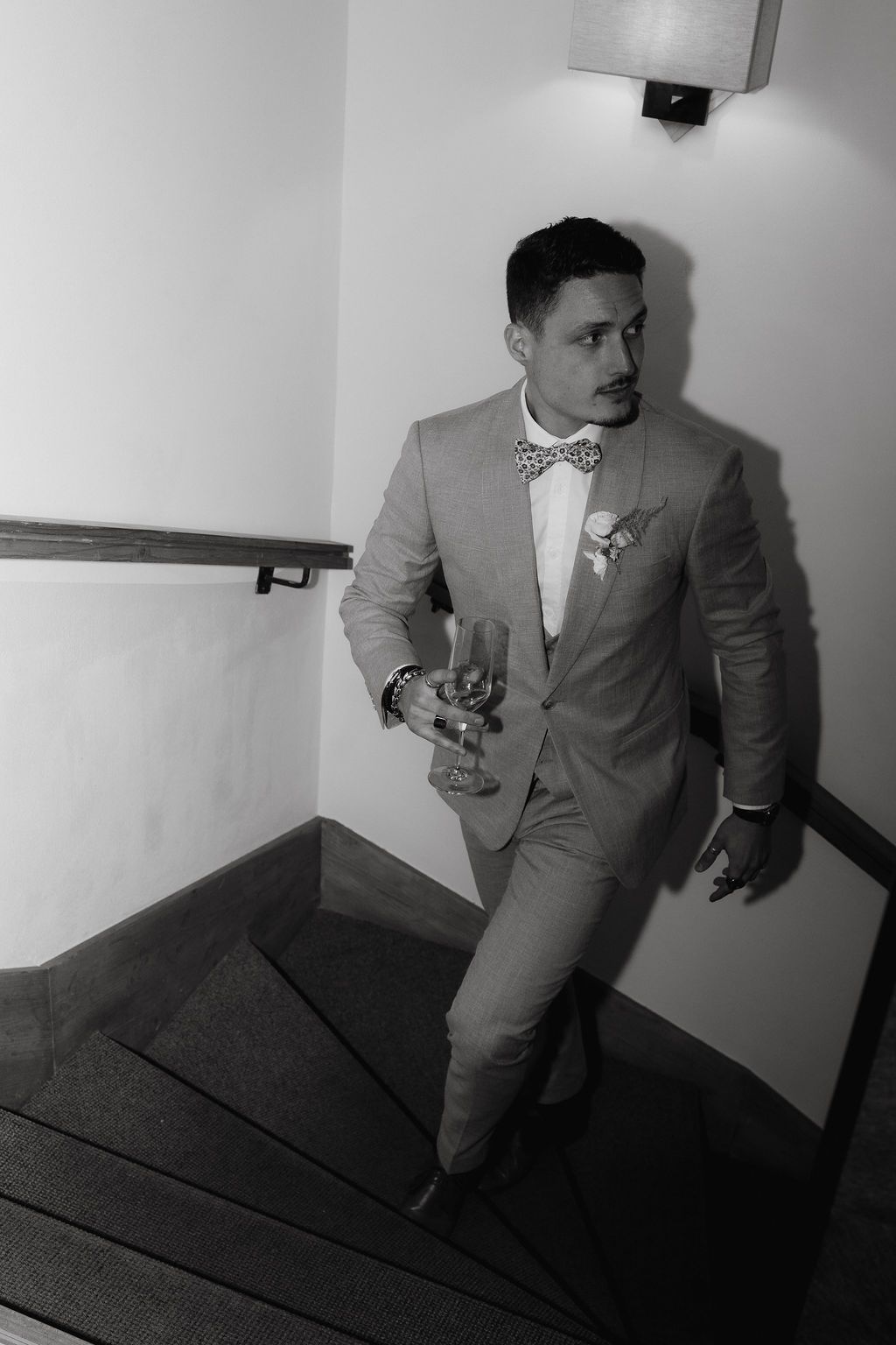 Elegant groom in gray suit and bow tie holds drink while descending staircase in black and white photo.