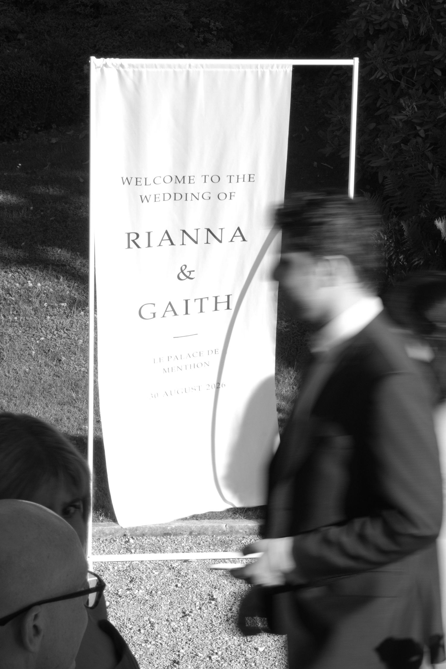 Black and white photo of a wedding welcome sign for Rianna and Gaith with guests walking past.