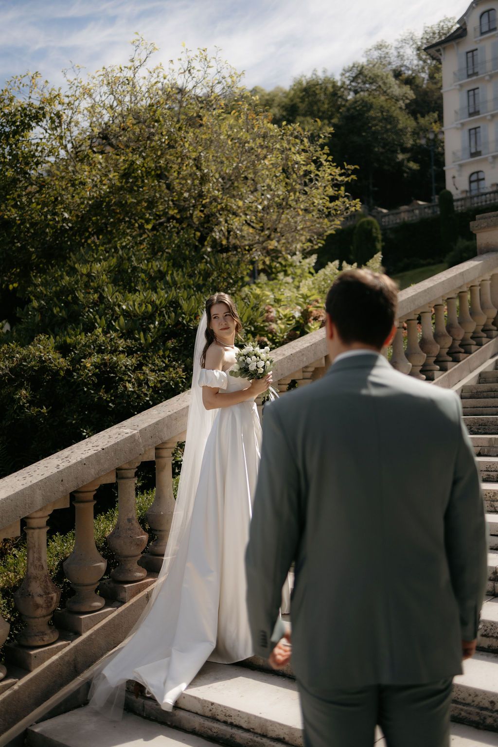 Bride in elegant white gown and veil smiles at groom on sunlit stone balustrade staircase.