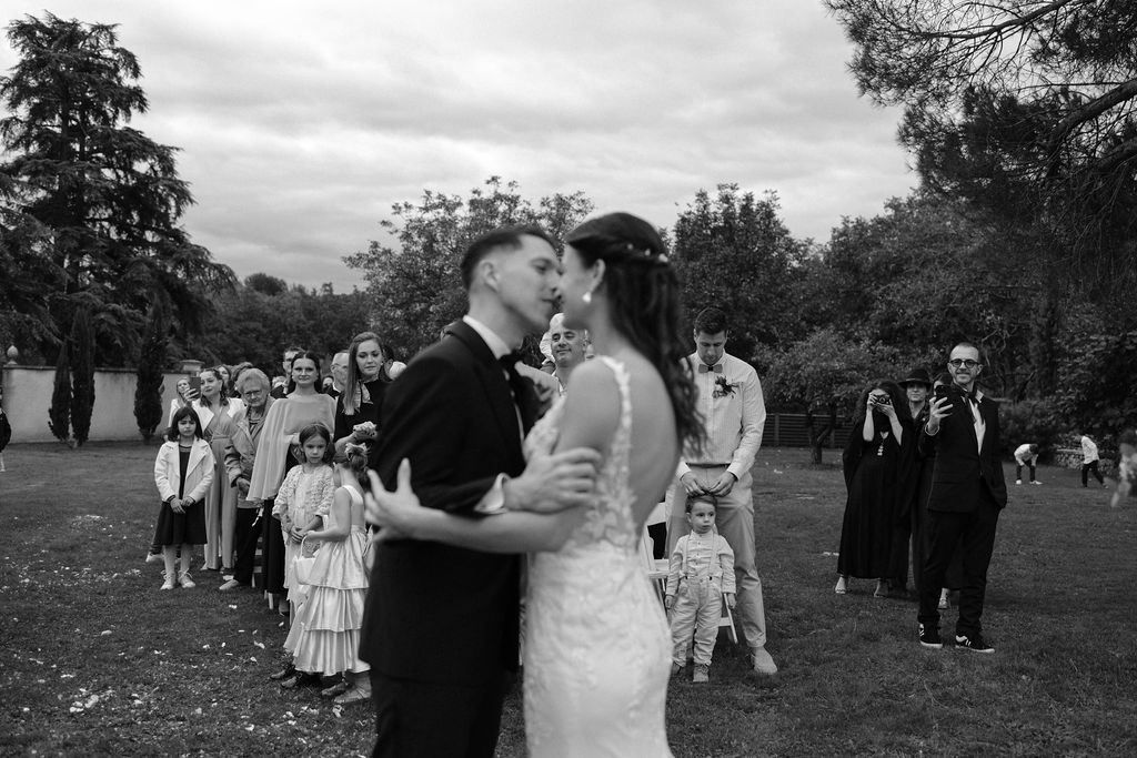 Black and white photo of bride and groom sharing first kiss surrounded by wedding guests outdoors.