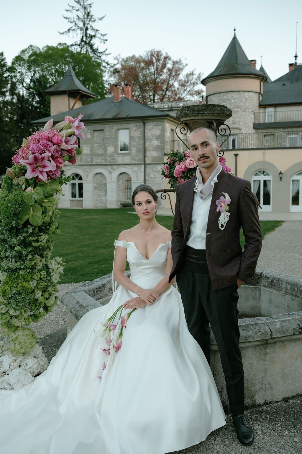 Bride and groom pose elegantly before a French chateau surrounded by pink floral arrangements at dusk.