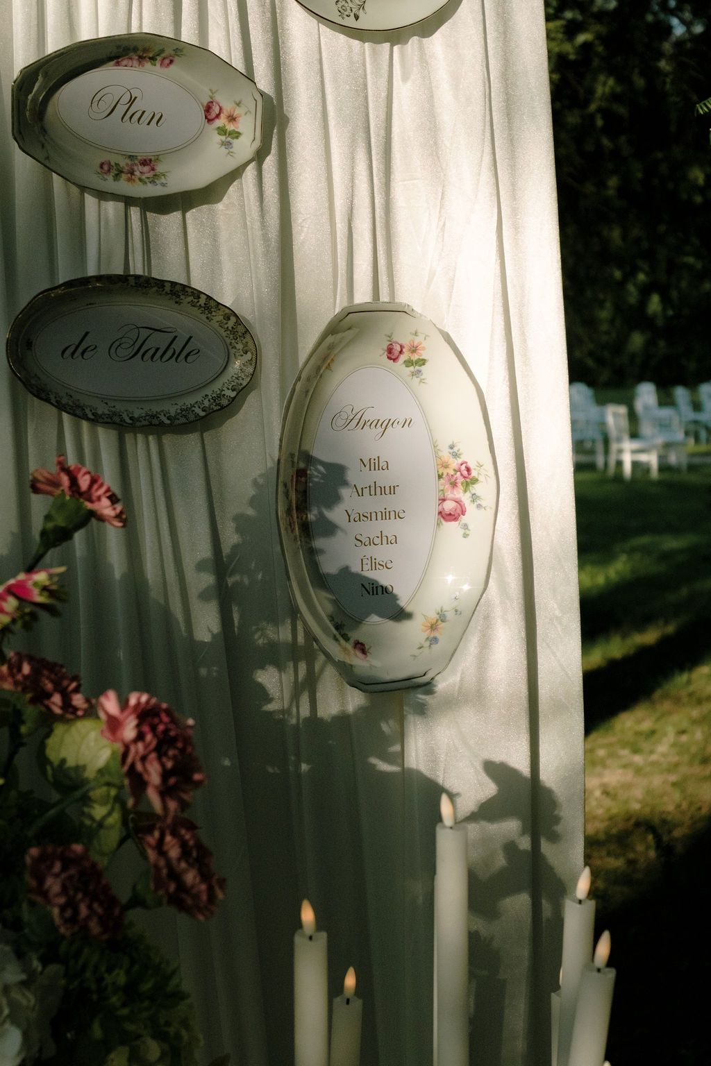 Outdoor wedding seating chart displayed on vintage floral porcelain plates with candles and flowers.