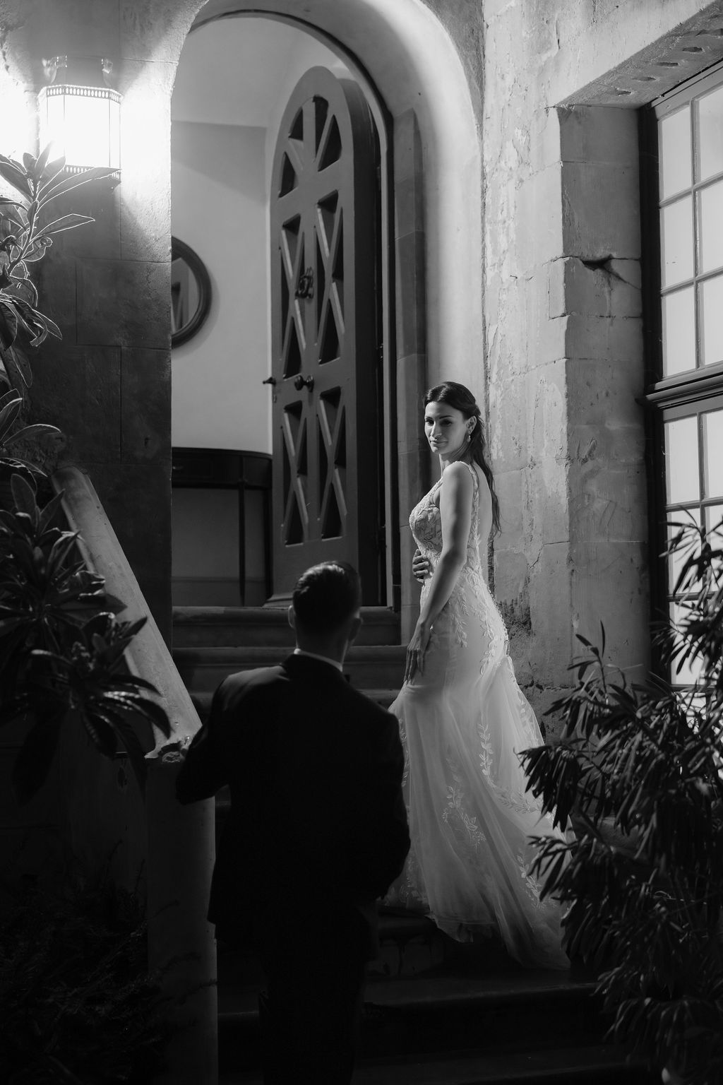 Black and white photo of bride in lace gown and groom at arched doorway of elegant venue.