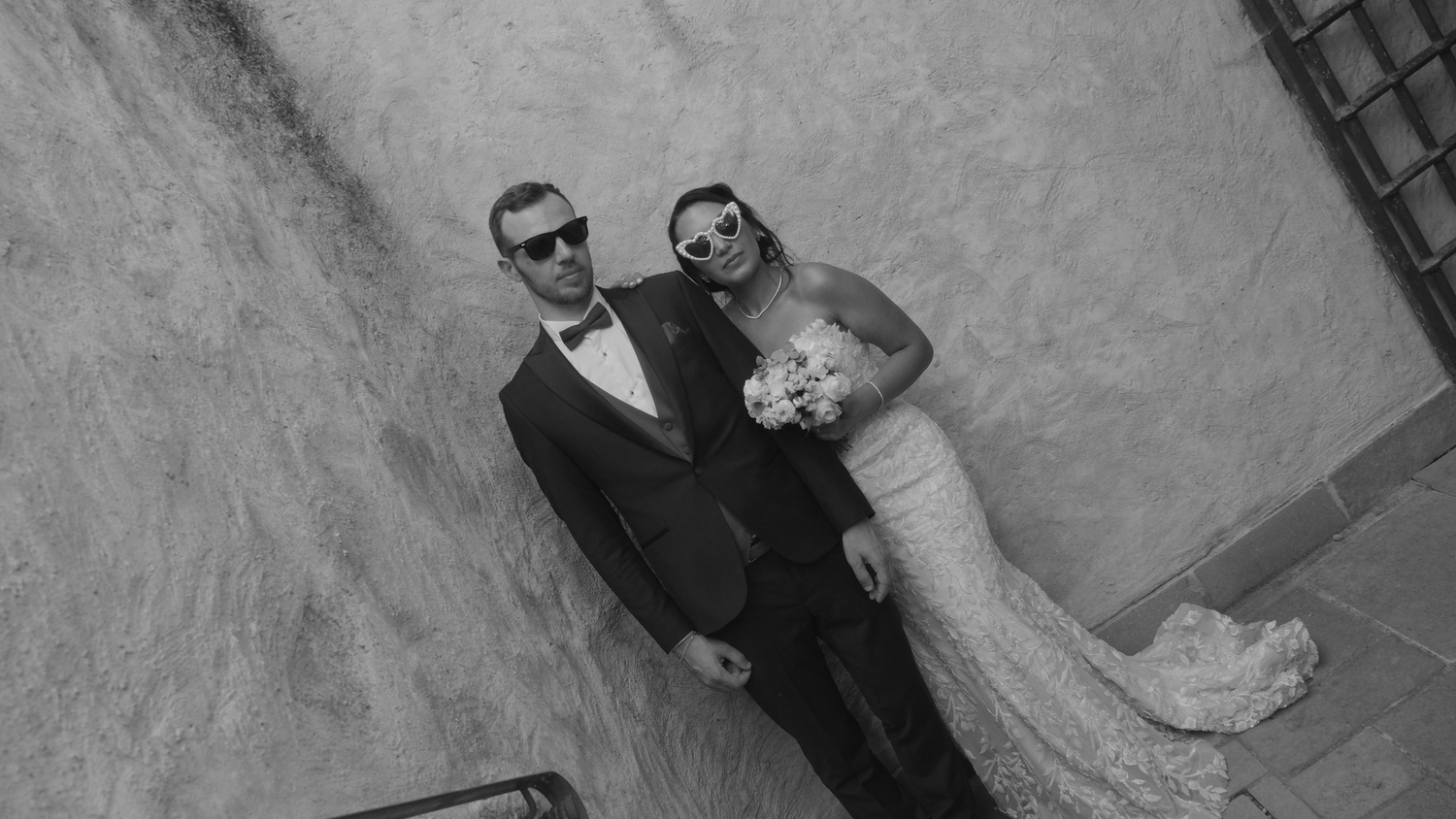 Black and white photo of a bride and groom in wedding attire, posing together against a textured wall.