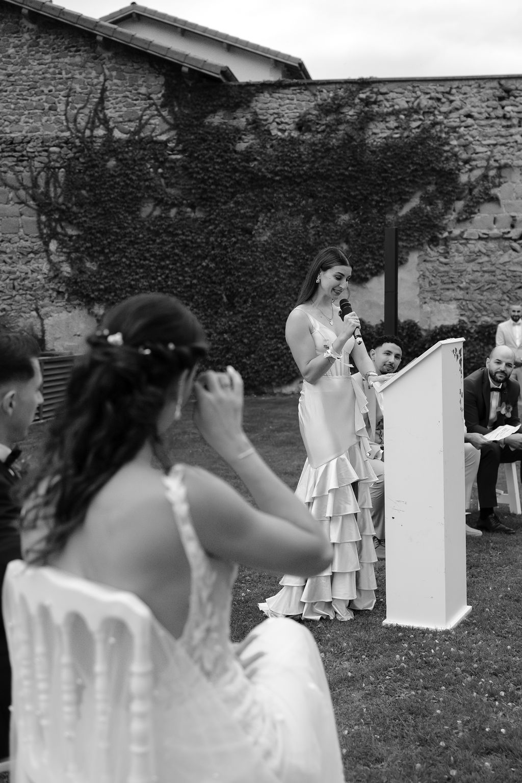 Black and white photo of woman in ruffled dress speaking at podium during outdoor wedding ceremony.