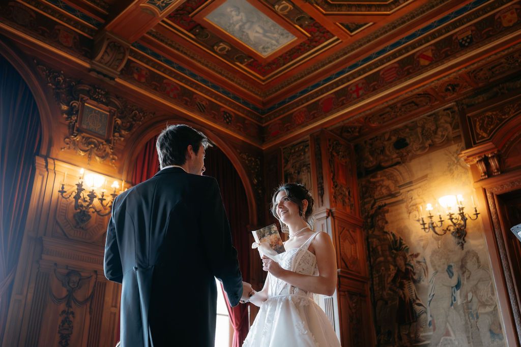 Bride and groom exchanging vows in ornately decorated room with painted ceiling and wood paneling.