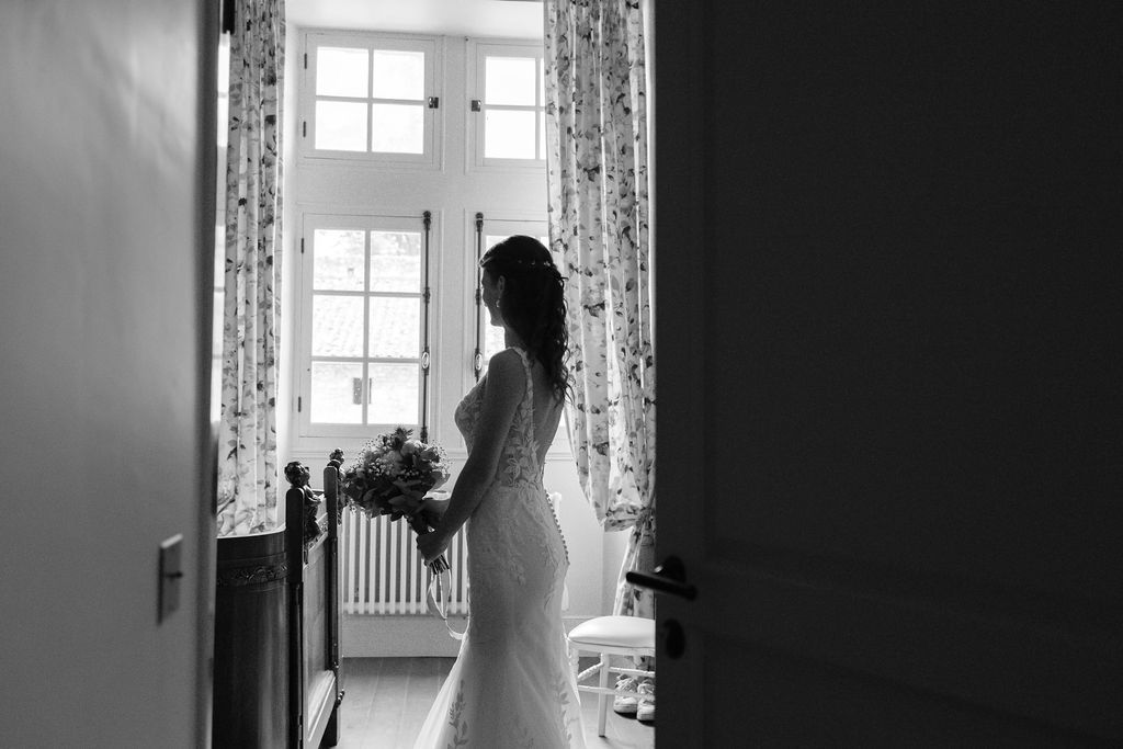 Bride in fitted lace wedding dress standing by window holding bouquet in black and white.