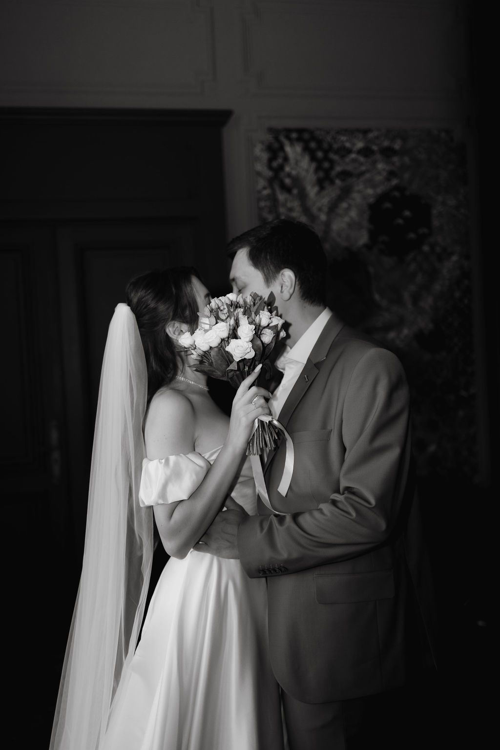 Black and white intimate portrait of bride and groom sharing kiss behind bridal bouquet of roses.