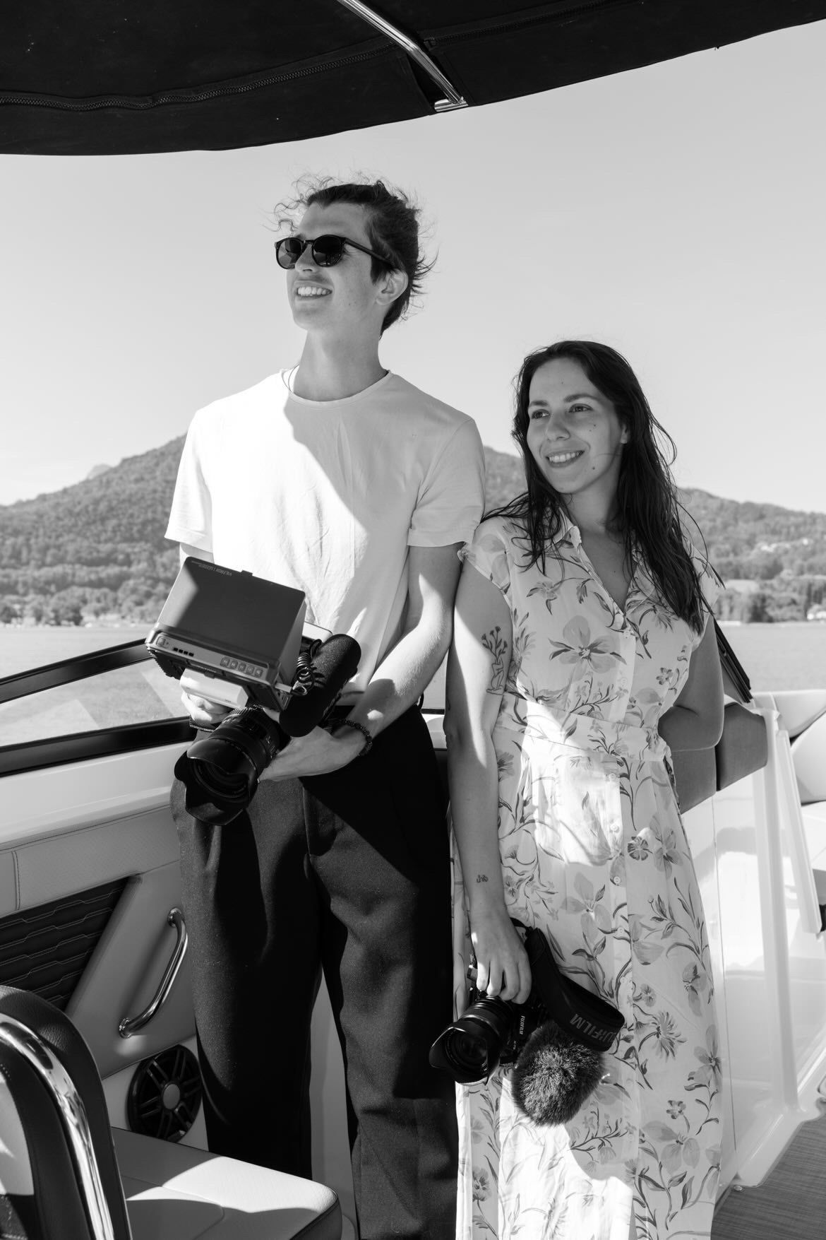 Two photographers holding cameras on a boat deck with scenic mountain views in black and white.