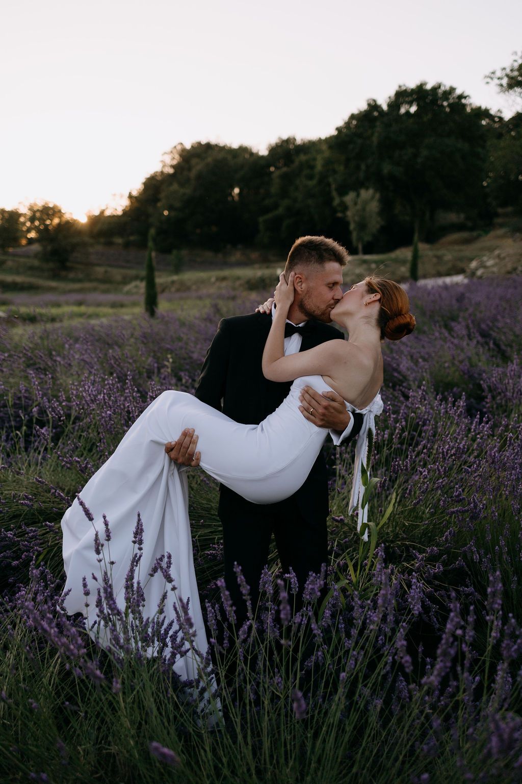 Groom dipping bride in white dress for a romantic kiss in a blooming lavender field at sunset.