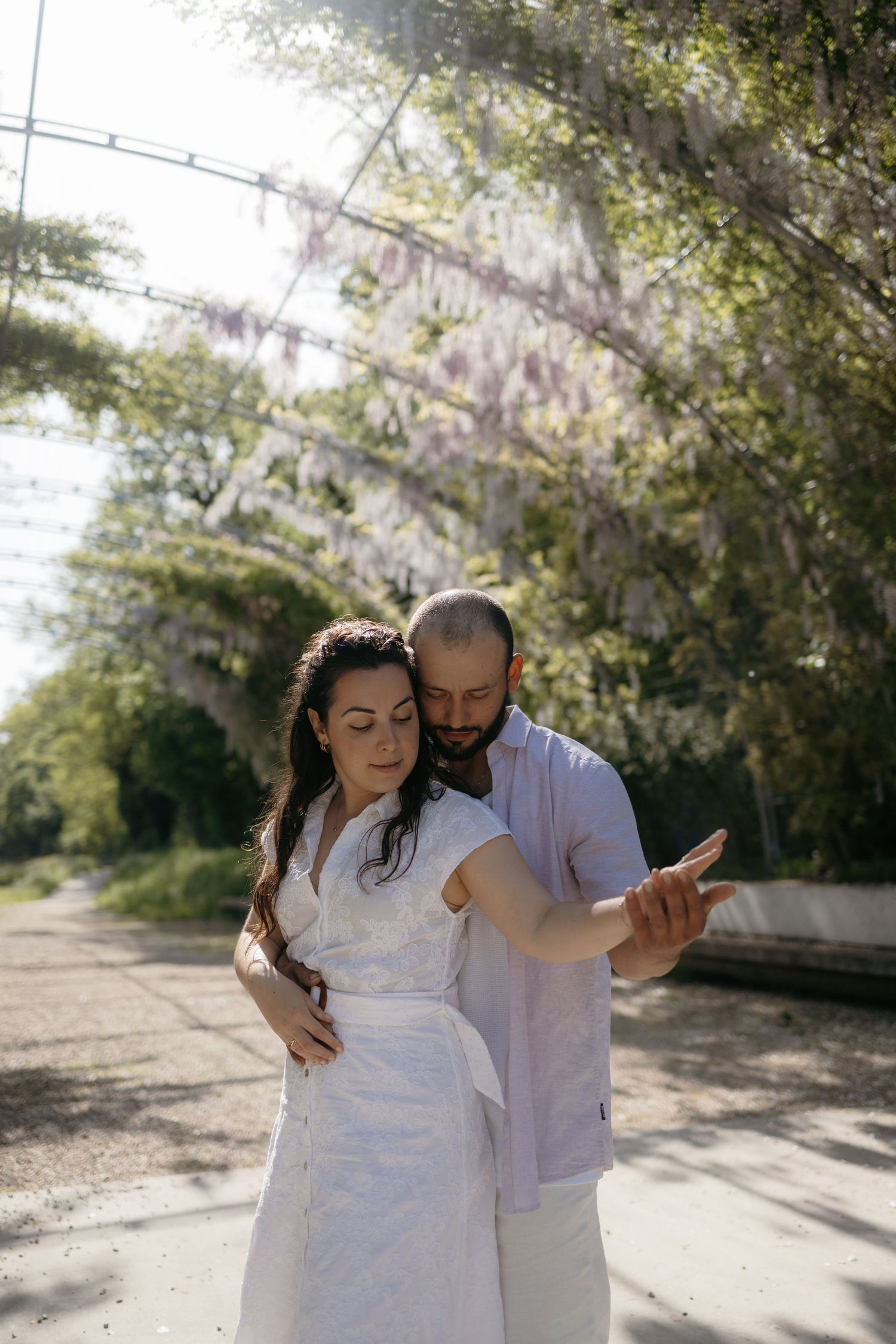 Couple in white attire embracing under a wisteria pergola in a sunlit garden path.