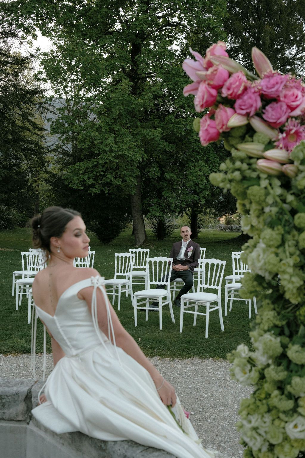 Bride in off-shoulder white gown sits on stone wall as groom waits among white chairs in lush garden ceremony.