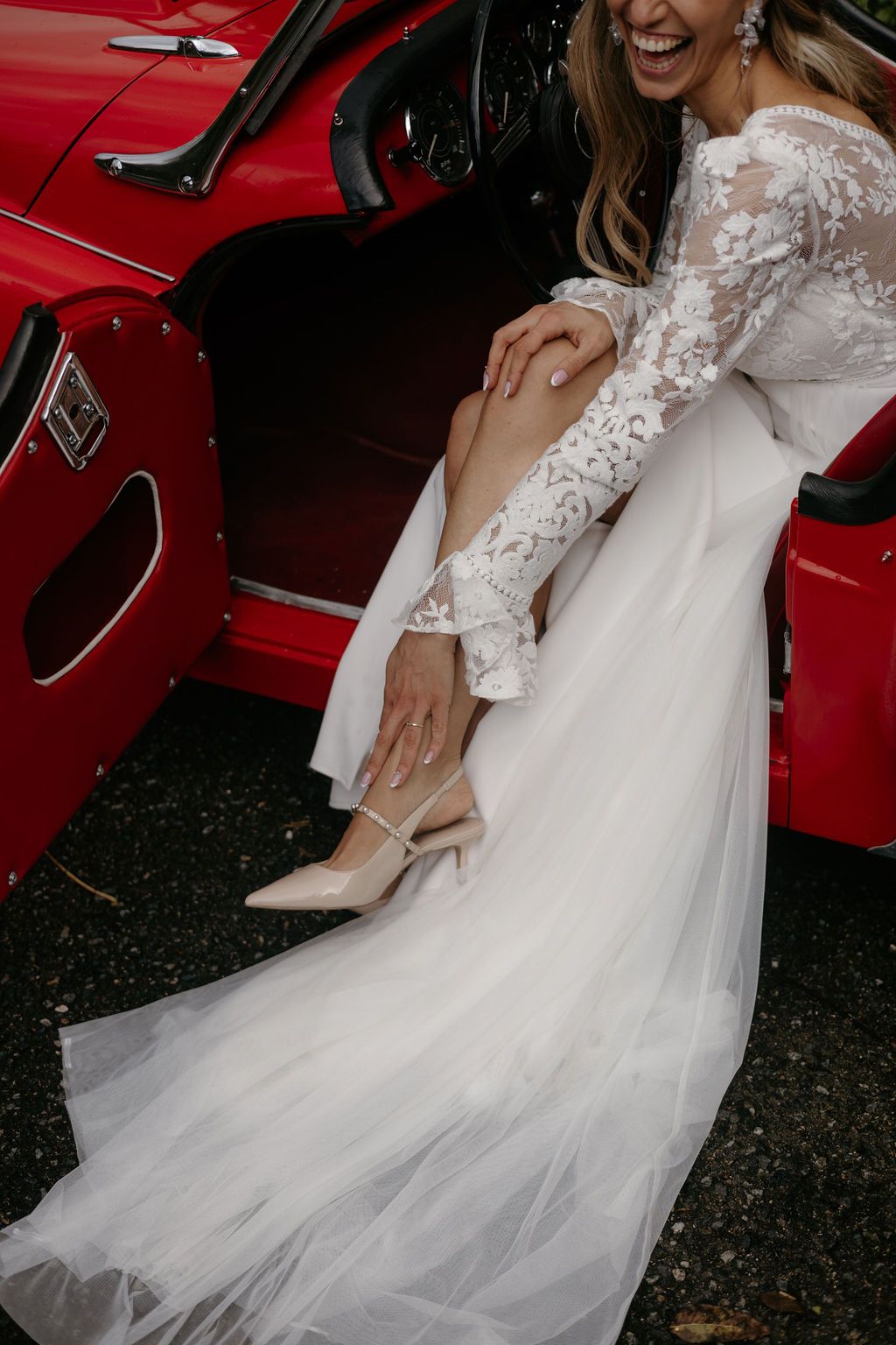 Joyful bride in long-sleeve lace gown exits red vintage convertible car on wedding day.
