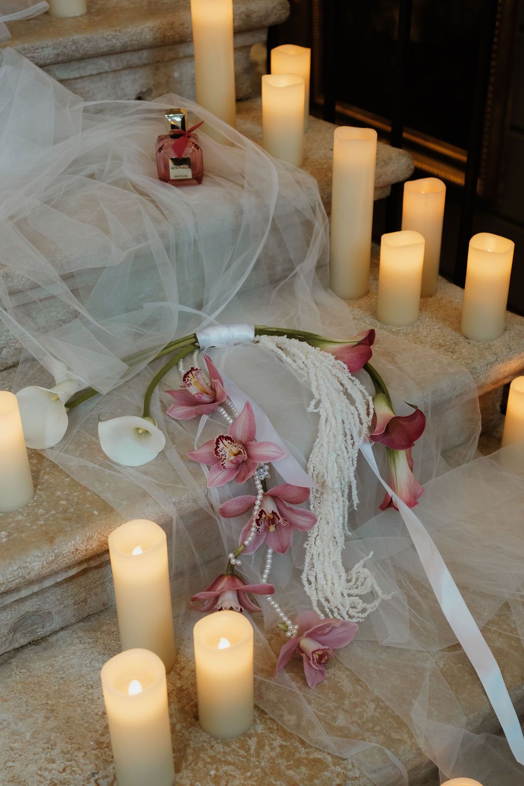 Elegant wedding decor with glowing pillar candles, pink orchids, pearls, and tulle on marble steps.