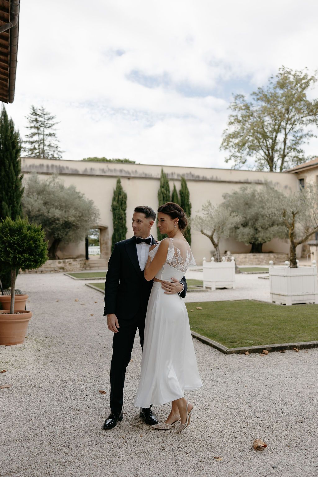 Bride and groom embracing in elegant courtyard with cypress trees and stone walls in Tuscany.